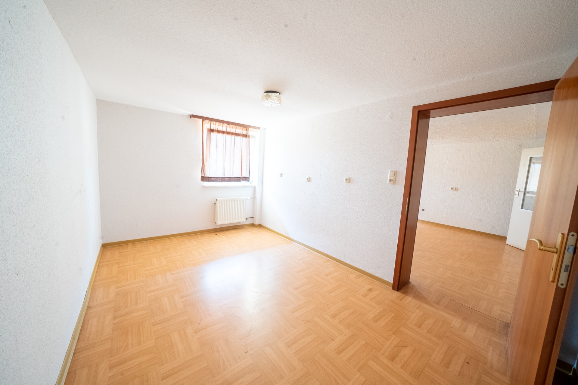 Leerer Raum mit Parkettboden, weiß gestrichenen Wänden und Fenster mit braunem Vorhang.