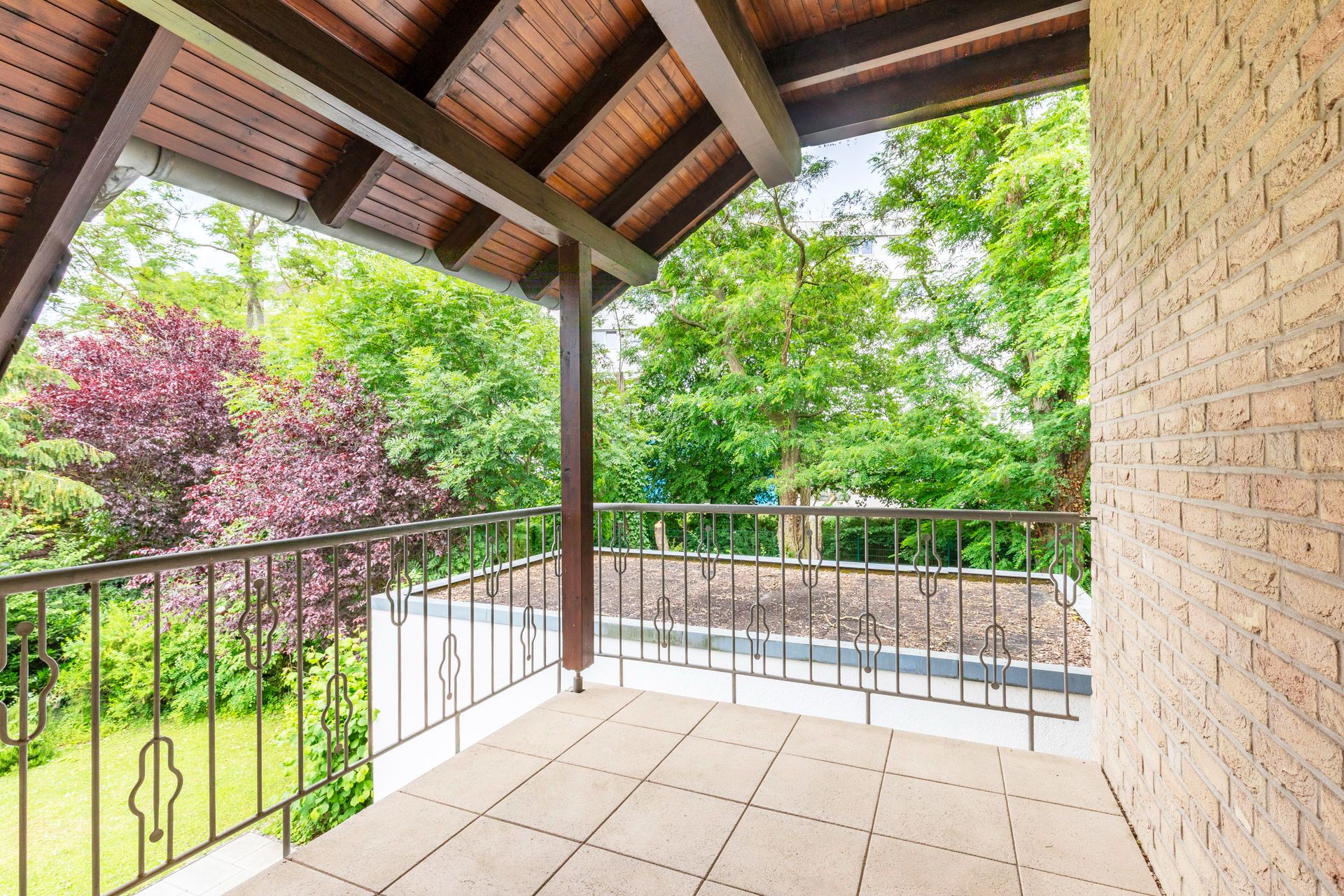 Überdachter Balkon mit Fliesenboden, Metallgeländer und Blick auf einen grünen Garten.