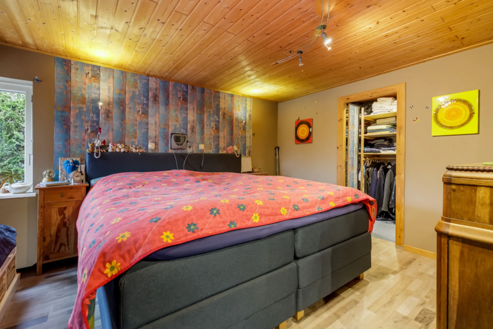 Schlafzimmer mit großem Bett, bunter Decke und offenem Kleiderschrank, Holzboden und Holzdecke.