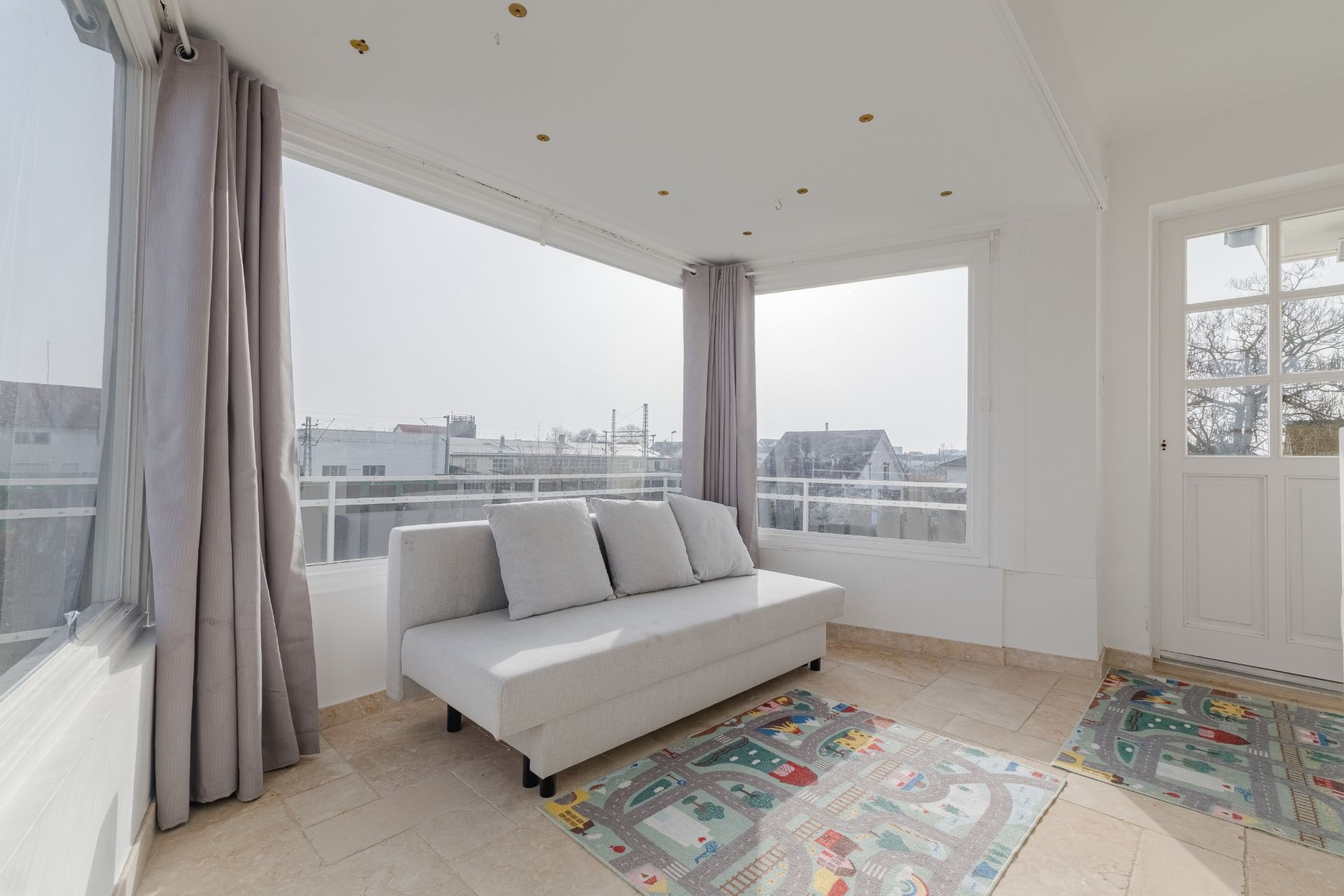 Heller Raum mit großem Sofa, großen Fenstern, Vorhängen und zwei Teppichen mit Straßenmotiven.