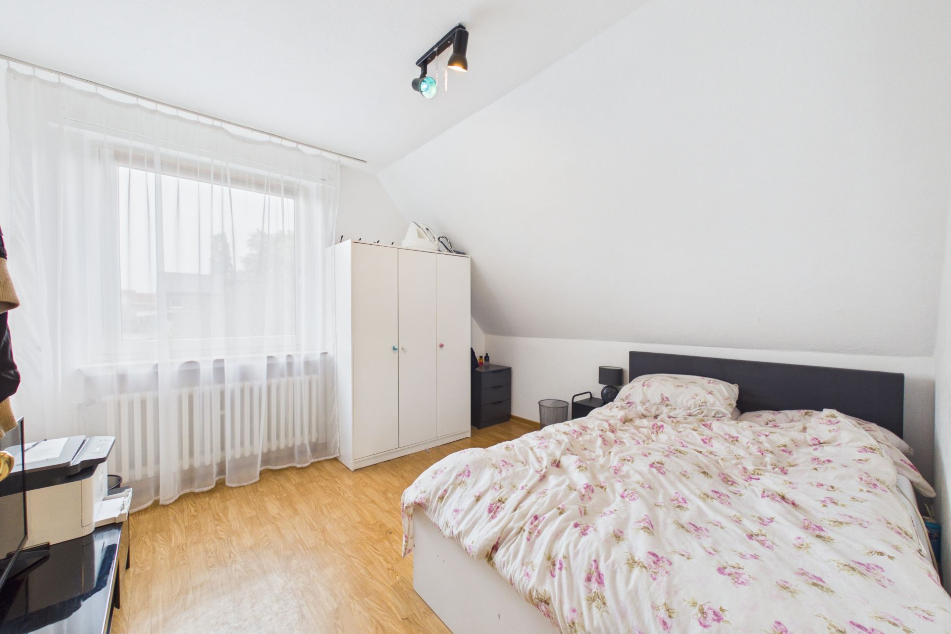 Helles Schlafzimmer mit Doppelbett, Kleiderschrank, Nachttischen und großem Fenster mit weißen Vorhängen.
