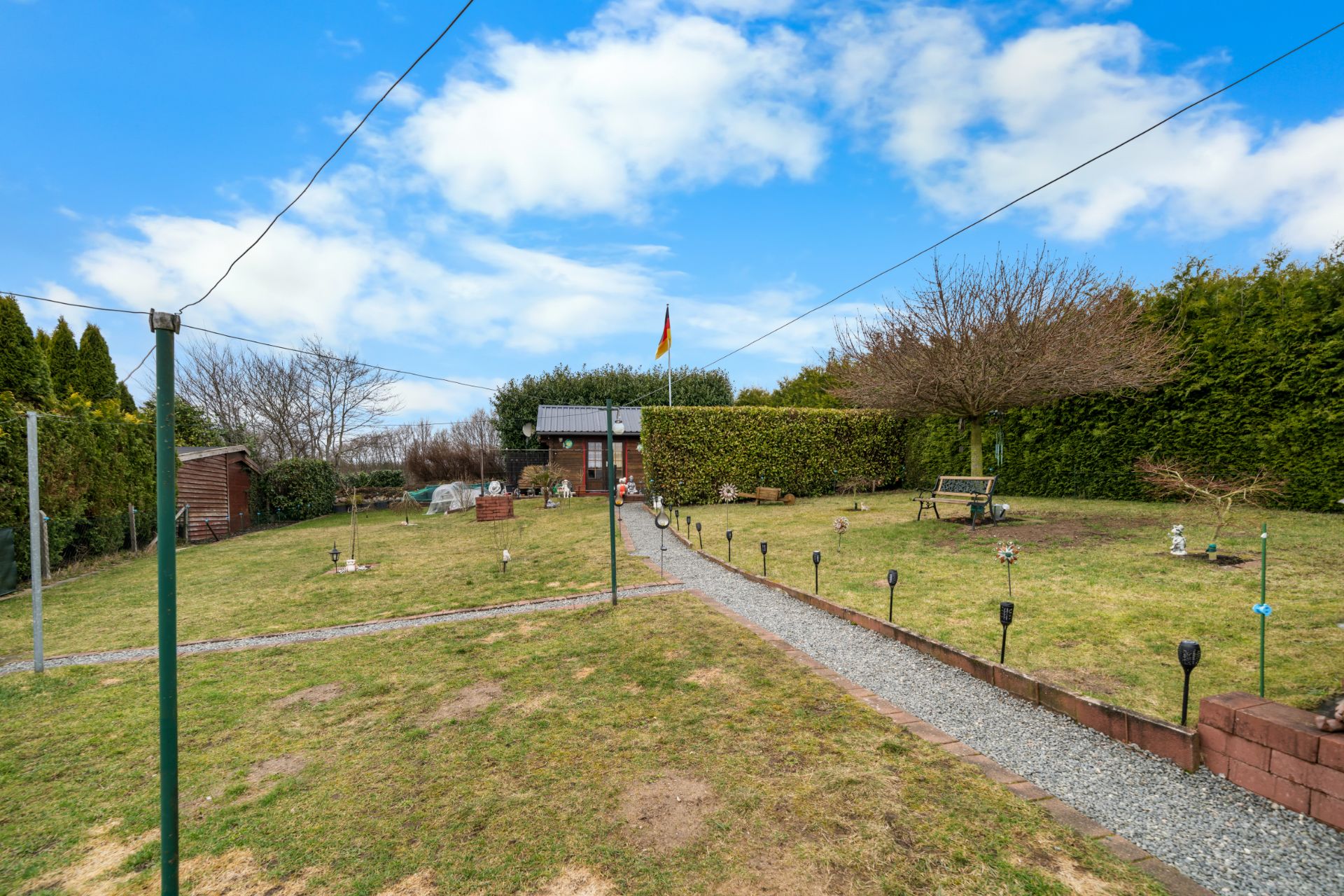 Garten mit Kiesweg, Sitzbank, Gartenhäuschen und deutscher Flagge bei blauem Himmel.