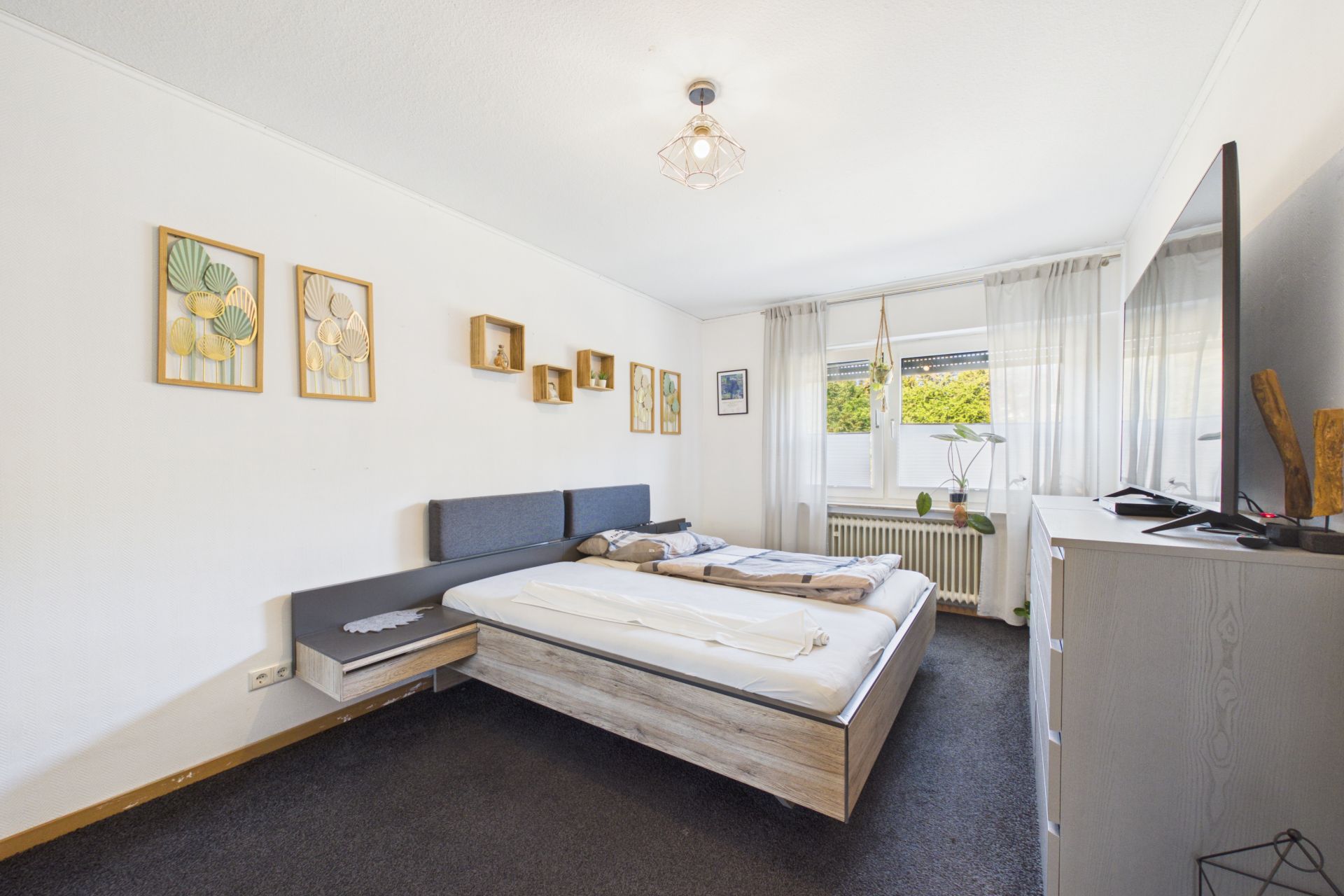 Helles Schlafzimmer mit Doppelbett, Kommode, Fernseher, Wanddeko und großem Fenster mit Gardinen.