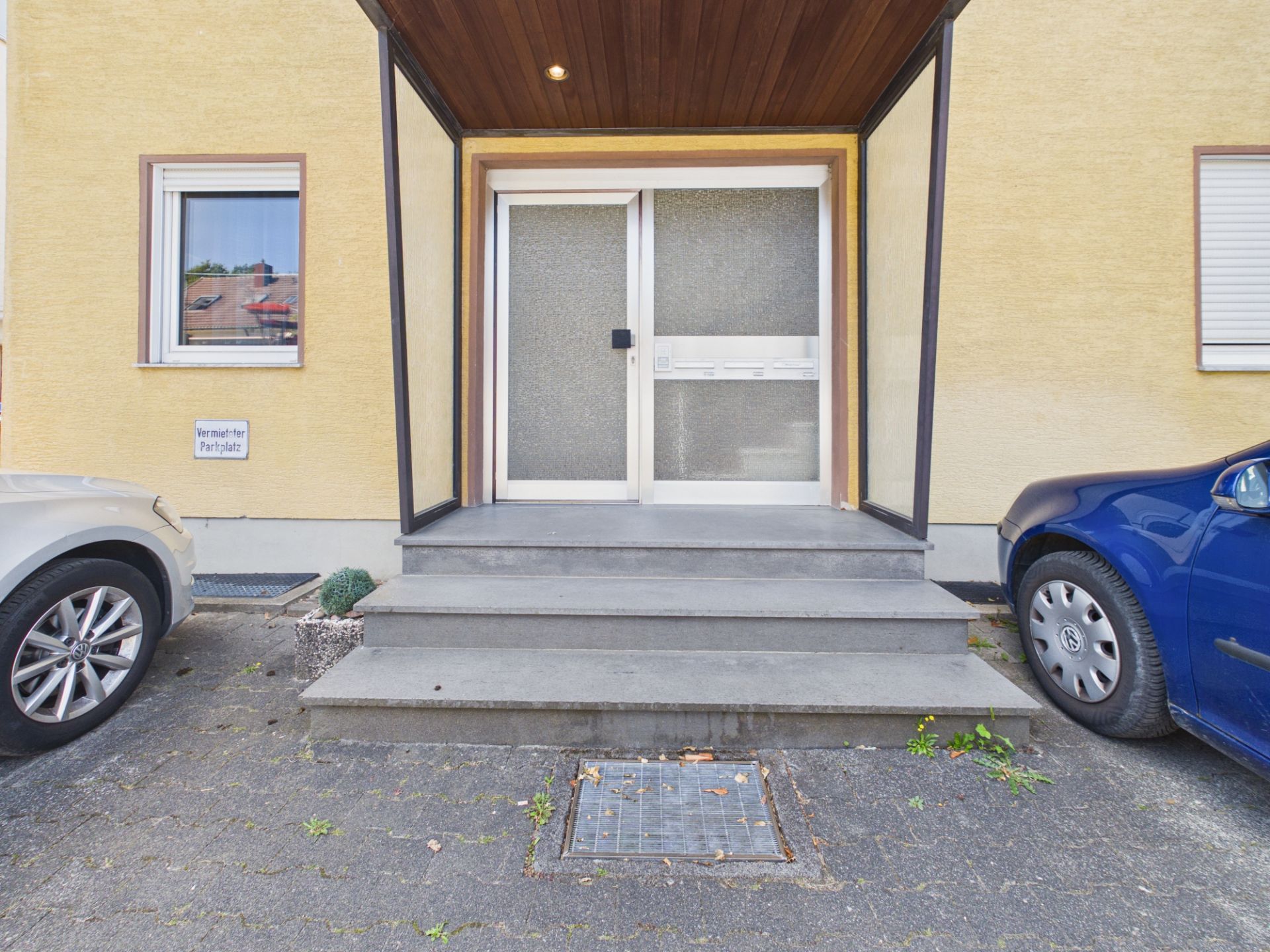 Eingangstür eines Wohnhauses mit Glas, Treppe und Autos links und rechts.