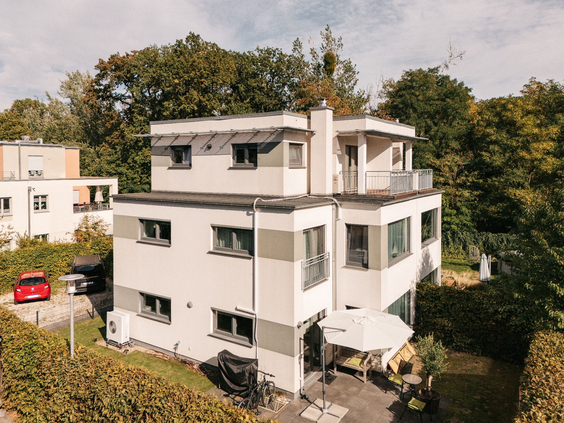 Modernes, mehrstöckiges Wohnhaus mit Terrasse, Sonnenschirm und Garten, umgeben von Hecken.