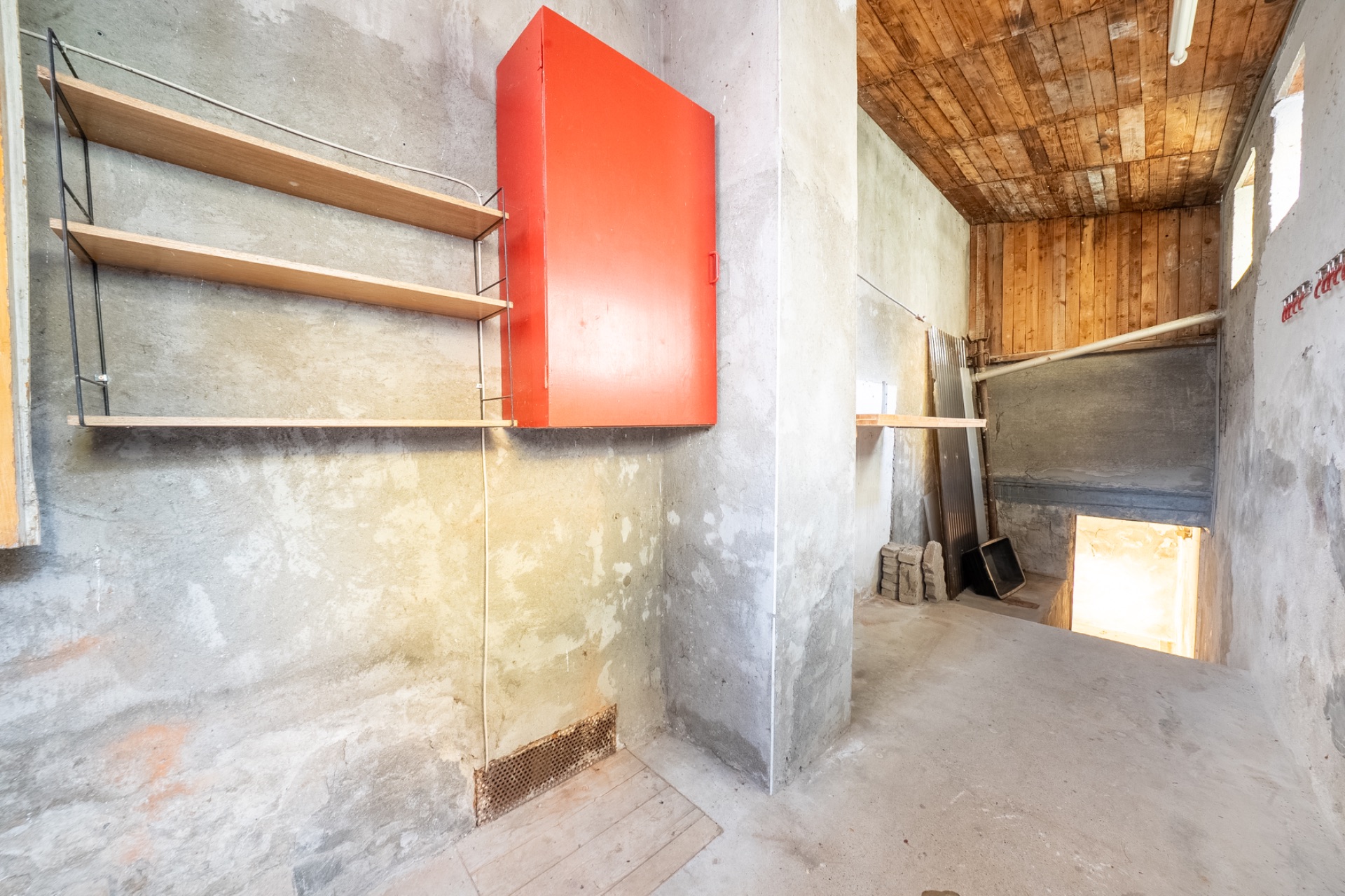 Ein schlichter Kellerraum mit Regalen, rotem Schrank und Holzdecke ist sichtbar.