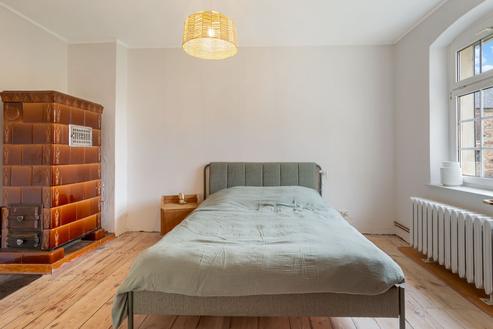 Schlafzimmer mit Bett, Nachttisch, großem Fenster, Ofen und Holzfußboden.