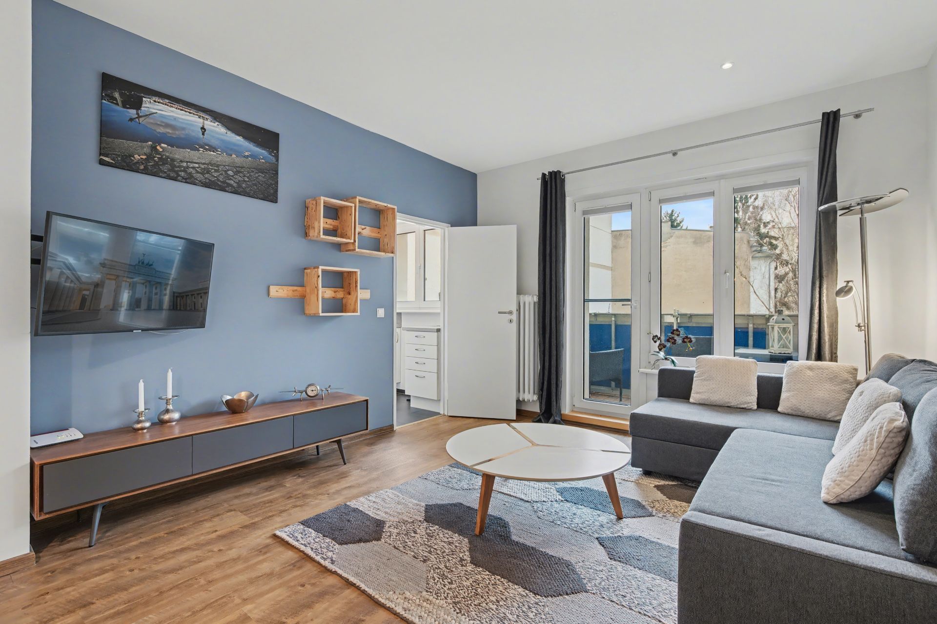 Modernes Wohnzimmer mit Ecksofa, Kaffeetisch, TV an blauer Wand und Zugang zum Balkon.