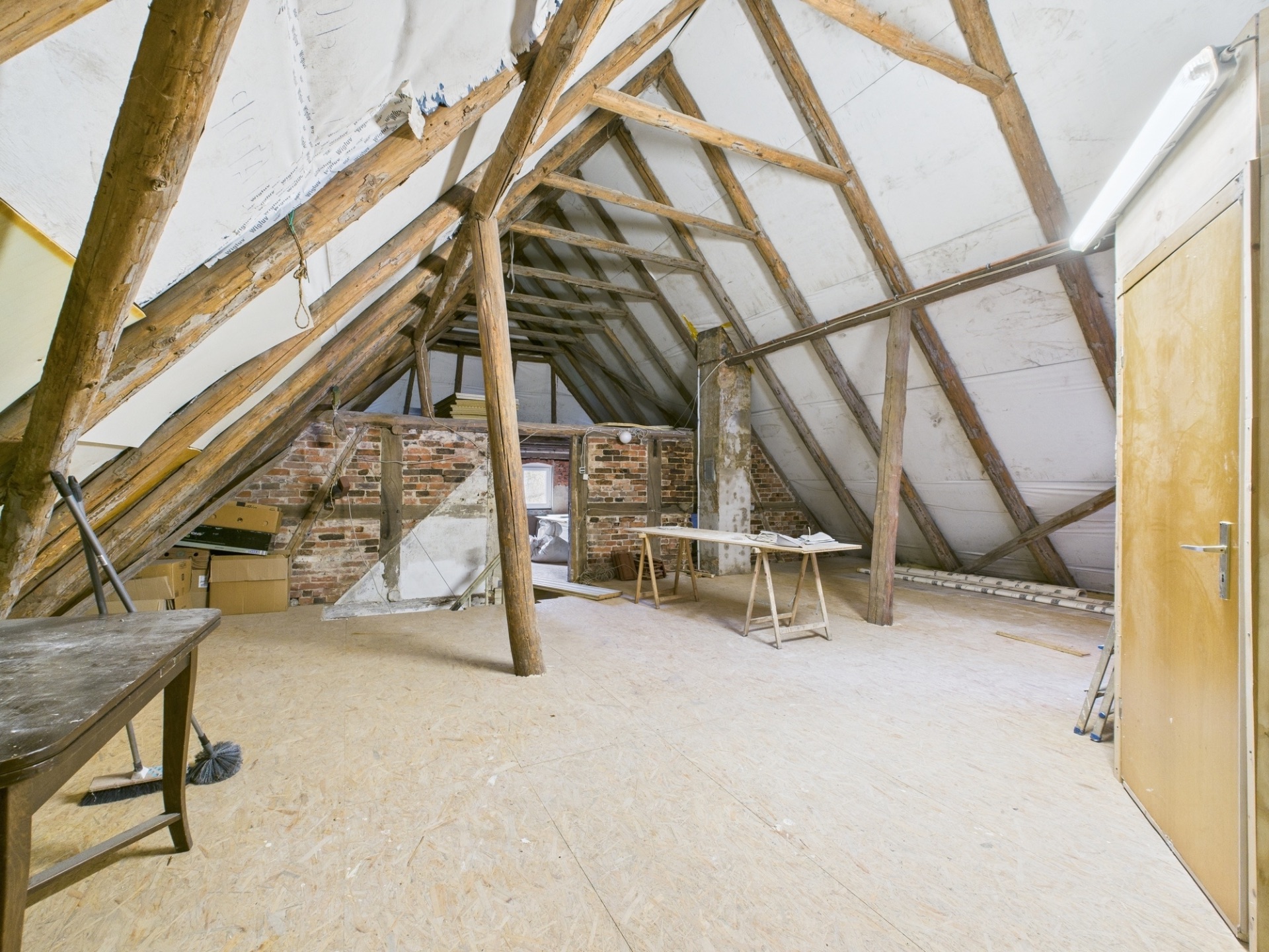 Heller Dachboden mit sichtbaren Holzbalken, Tisch, Kartons und Backsteinwand.