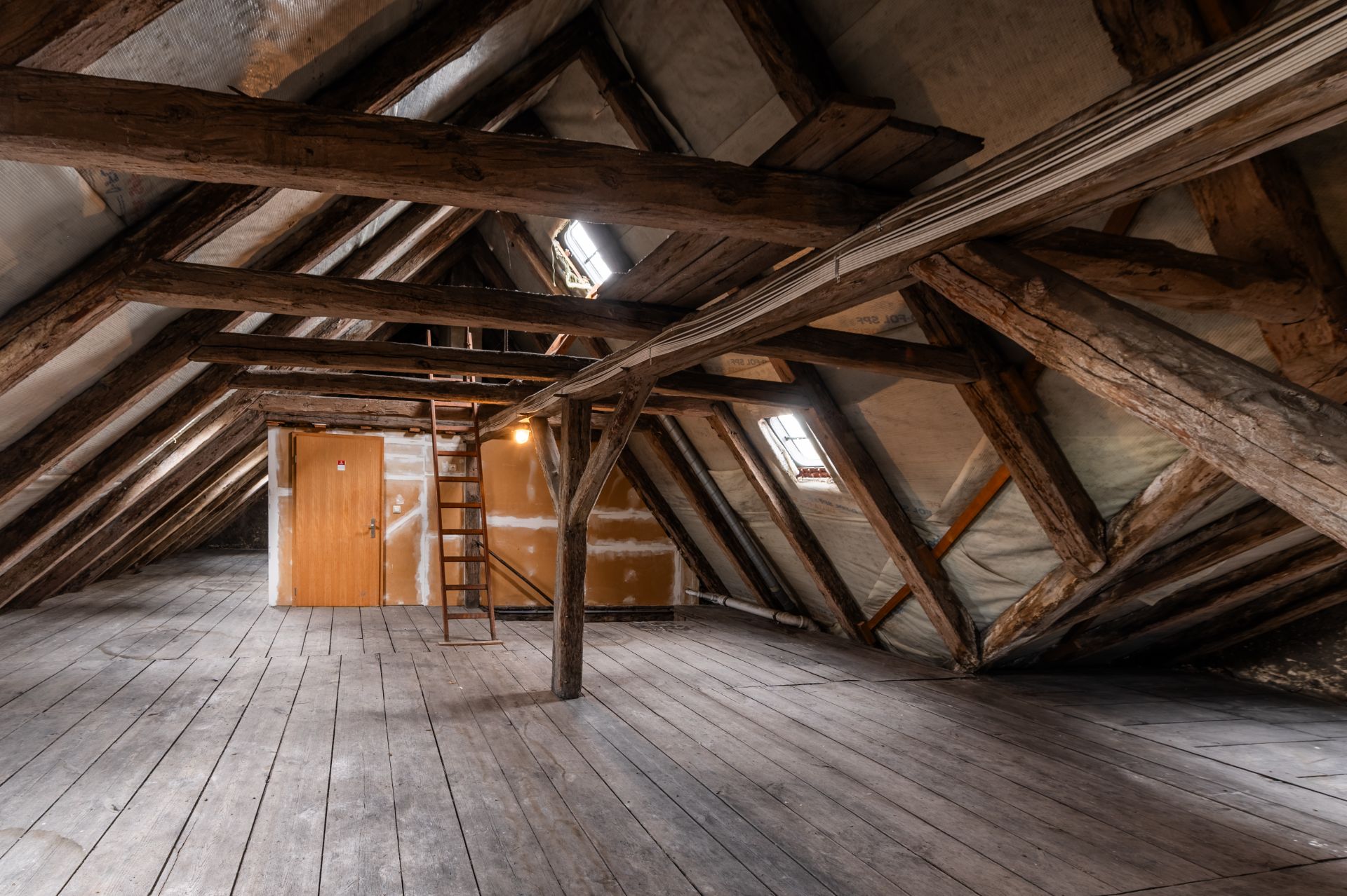 Dachbodenraum mit Holzbalken, Holztür, Leiter und kleinem Fenster.