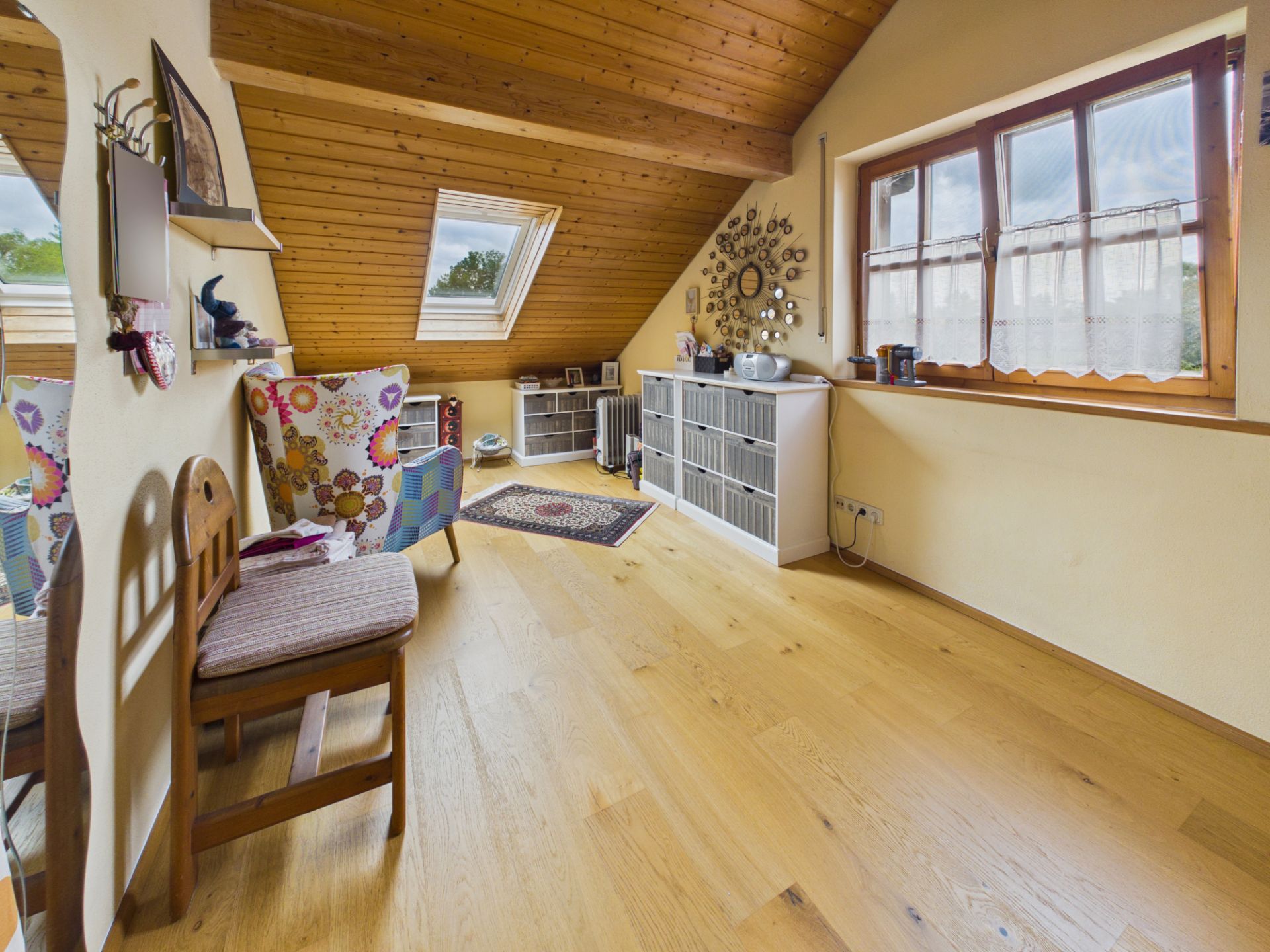 Helles Dachzimmer mit Holzdecke, Sessel, Stuhl und Regal, großes Fenster und Teppich.