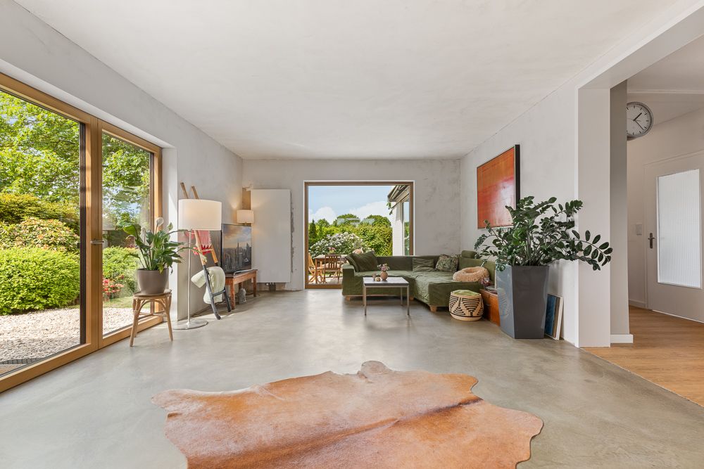 Helles Wohnzimmer mit großen Fenstern, Pflanzen, grünem Sofa und Teppich auf Betonboden.
