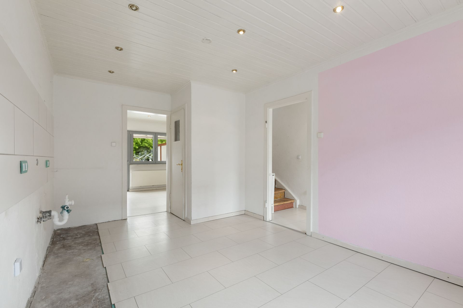 Leerer Raum mit Fliesenboden, rosa Wand und offenem Zugang zu Treppe und Fenster.