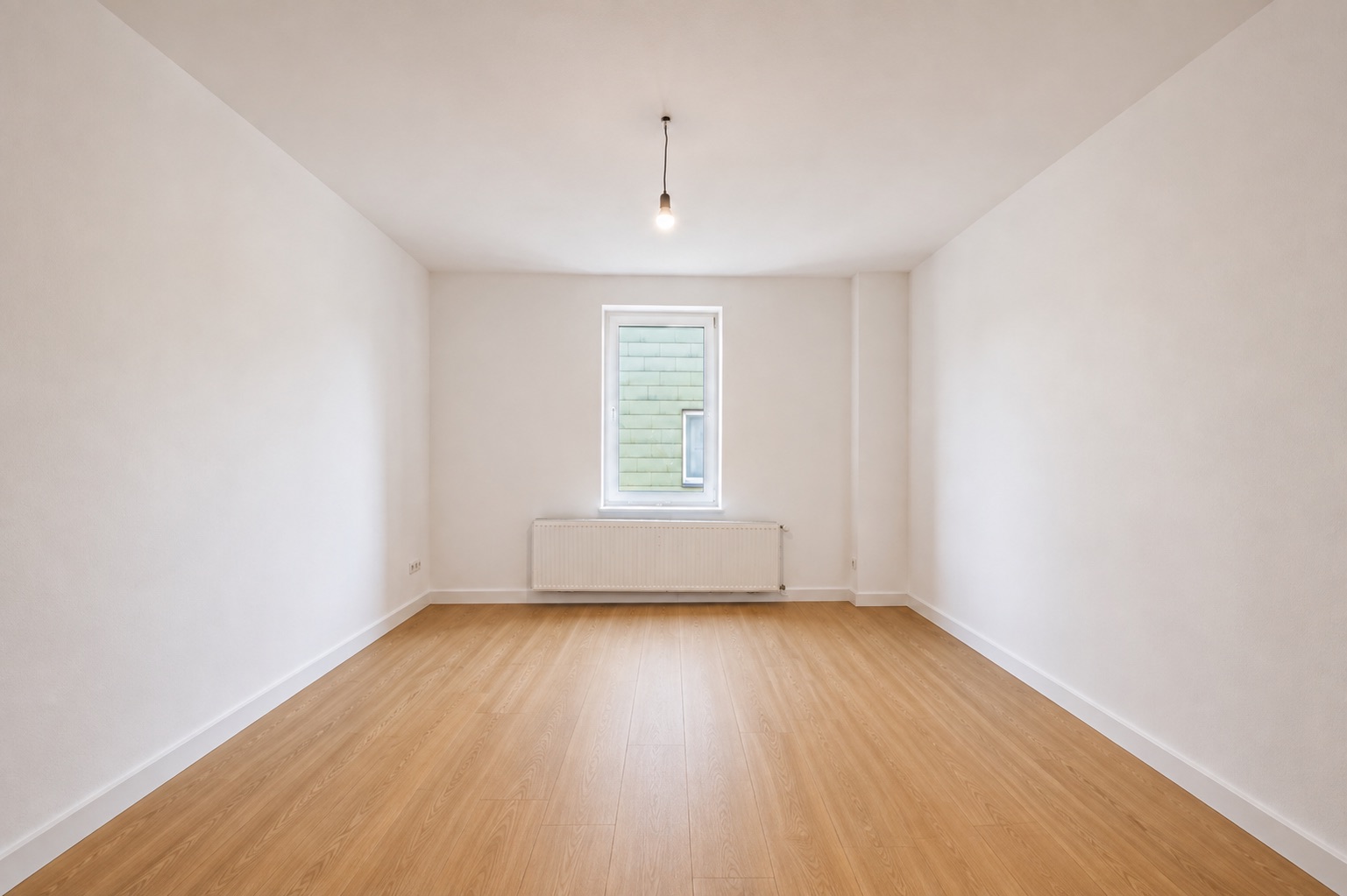 Leerer Raum mit hellem Holzboden, weißen Wänden, Fenster und Heizung an der Stirnseite.