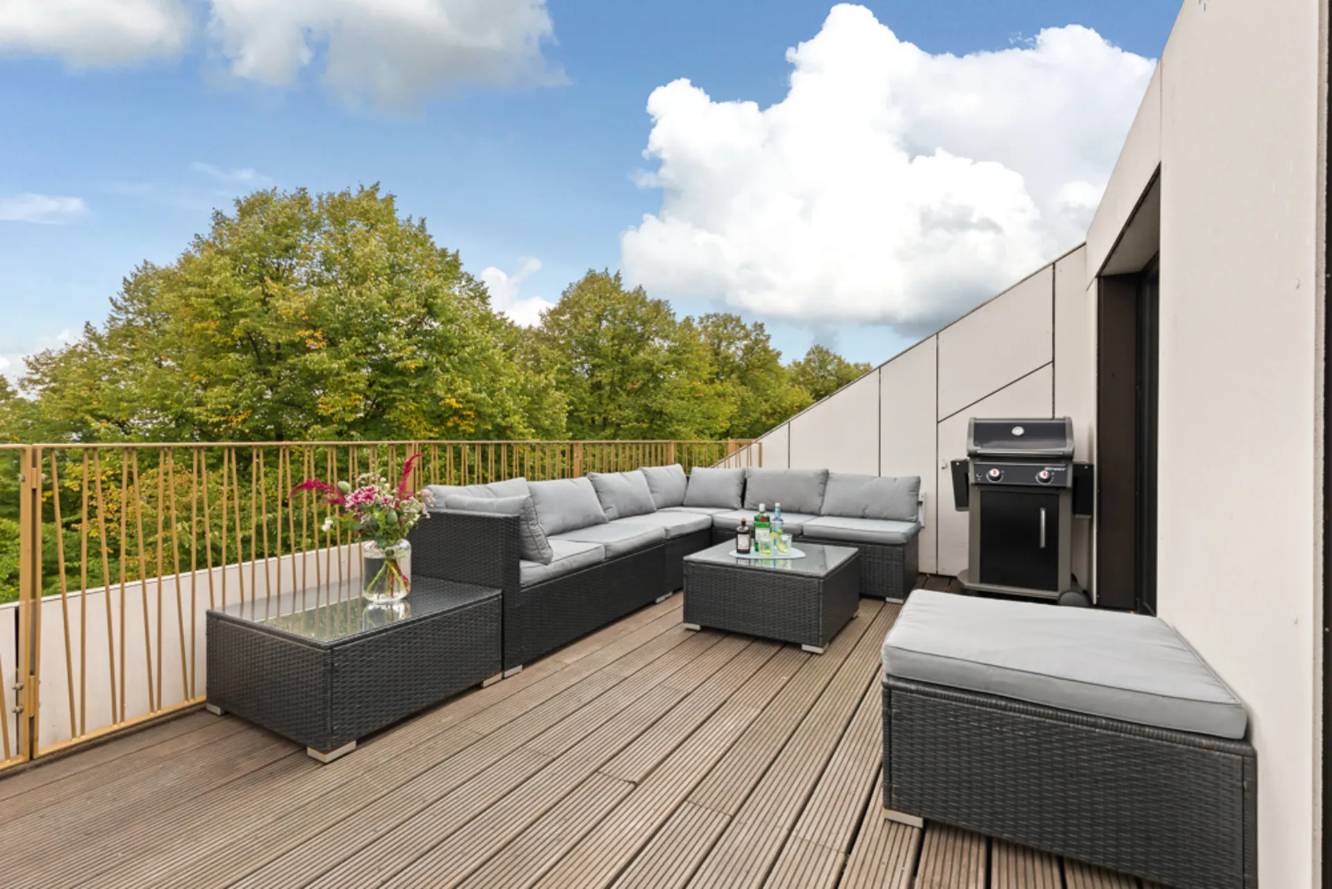 Große Dachterrasse mit Sofa, Tisch, Grill und Ausblick auf grüne Bäume.