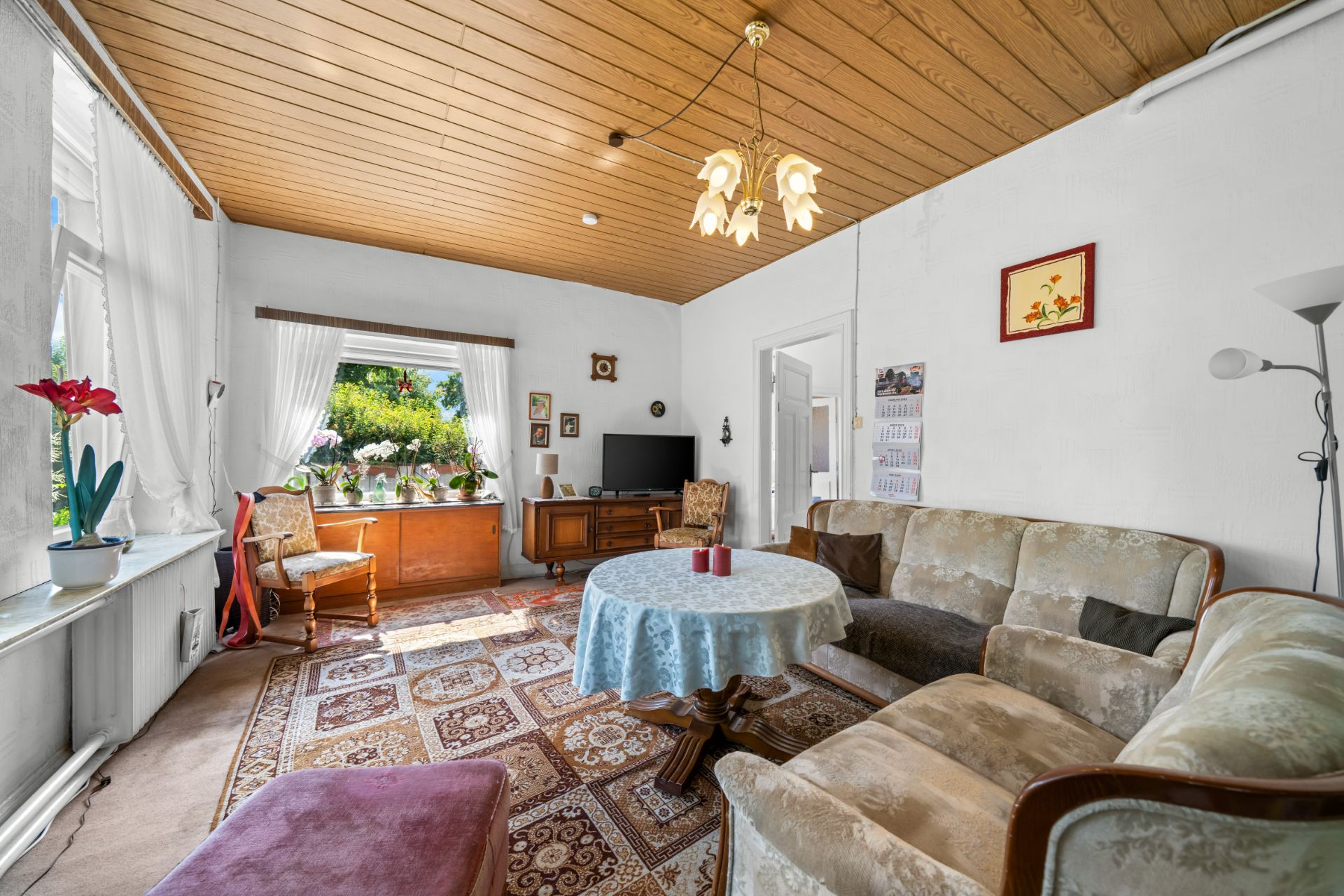 Helles Wohnzimmer mit Teppich, Sofa, Tisch, Fernseher, Sesseln und großen Fenstern mit Vorhängen.