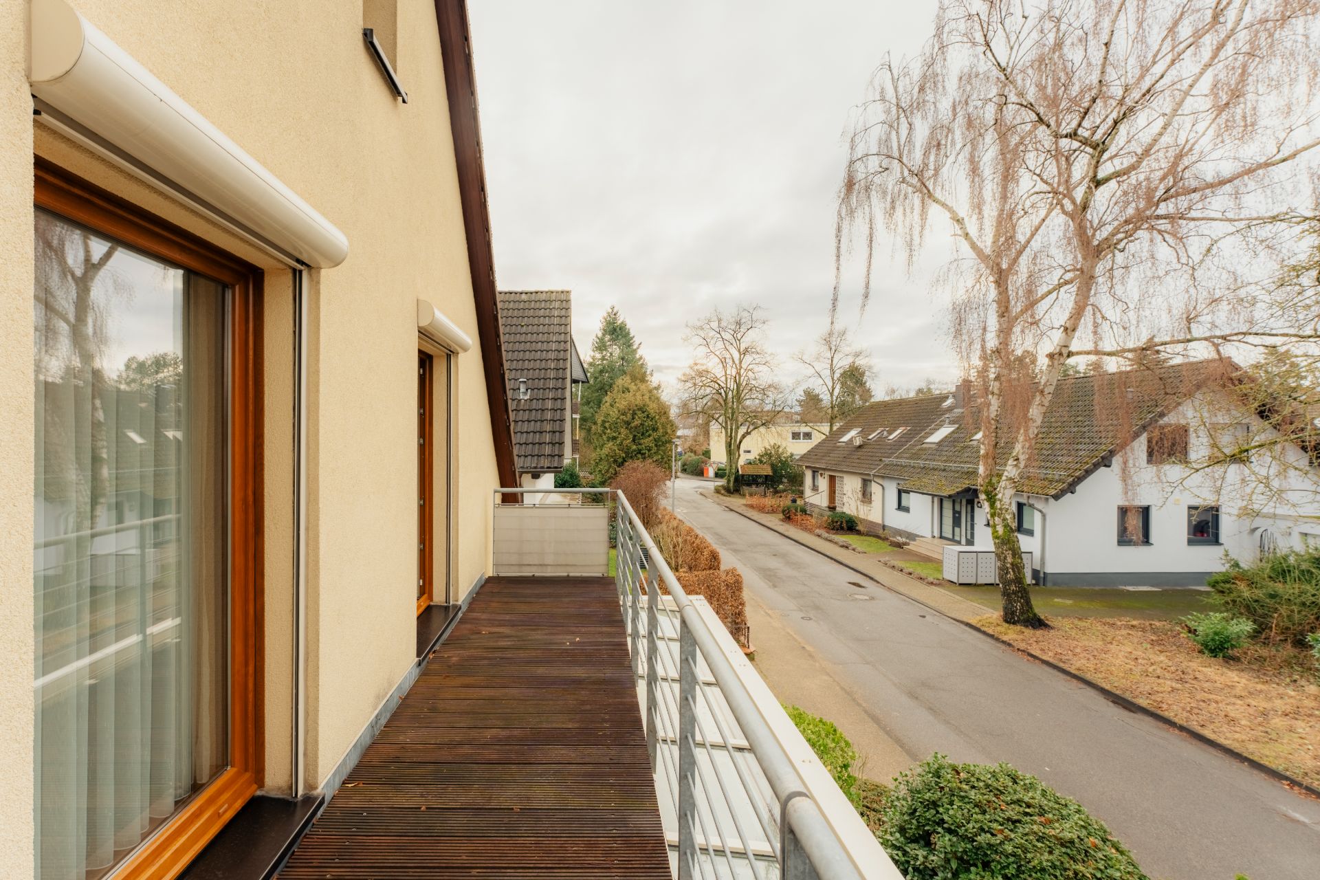 Balkon mit Holzboden an einem Wohnhaus, Blick auf eine ruhige Straße mit Bäumen und Häusern.