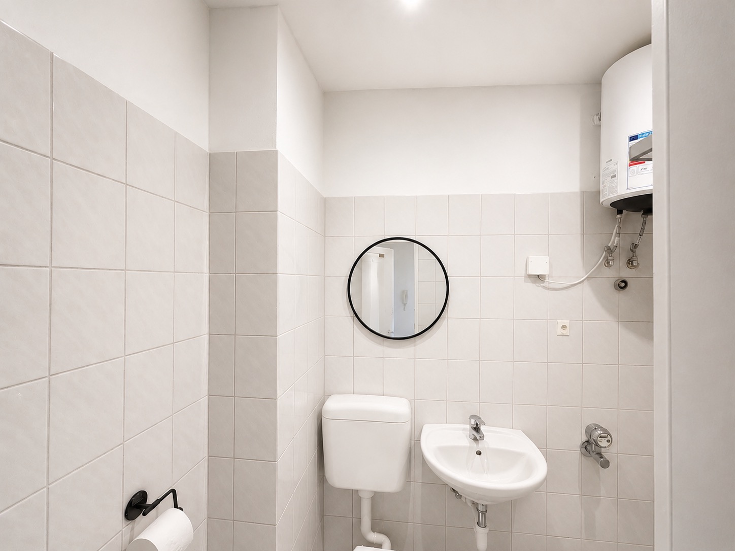 Helles Bad mit WC, Waschbecken, rundem Spiegel und Boiler an der Wand.