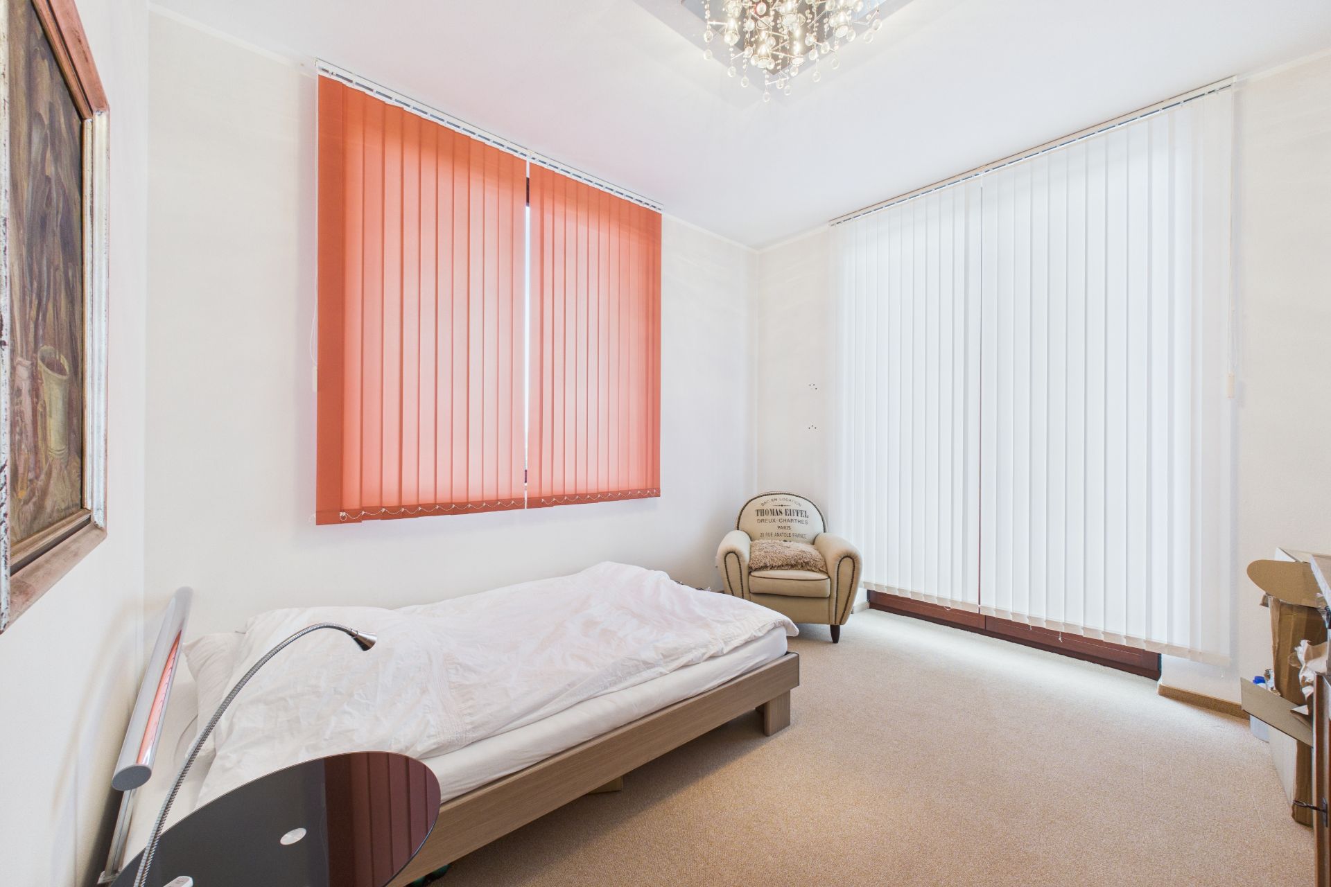 Helles Schlafzimmer mit Einzelbett, Sessel, großen Fenstern und vertikalen Jalousien.
