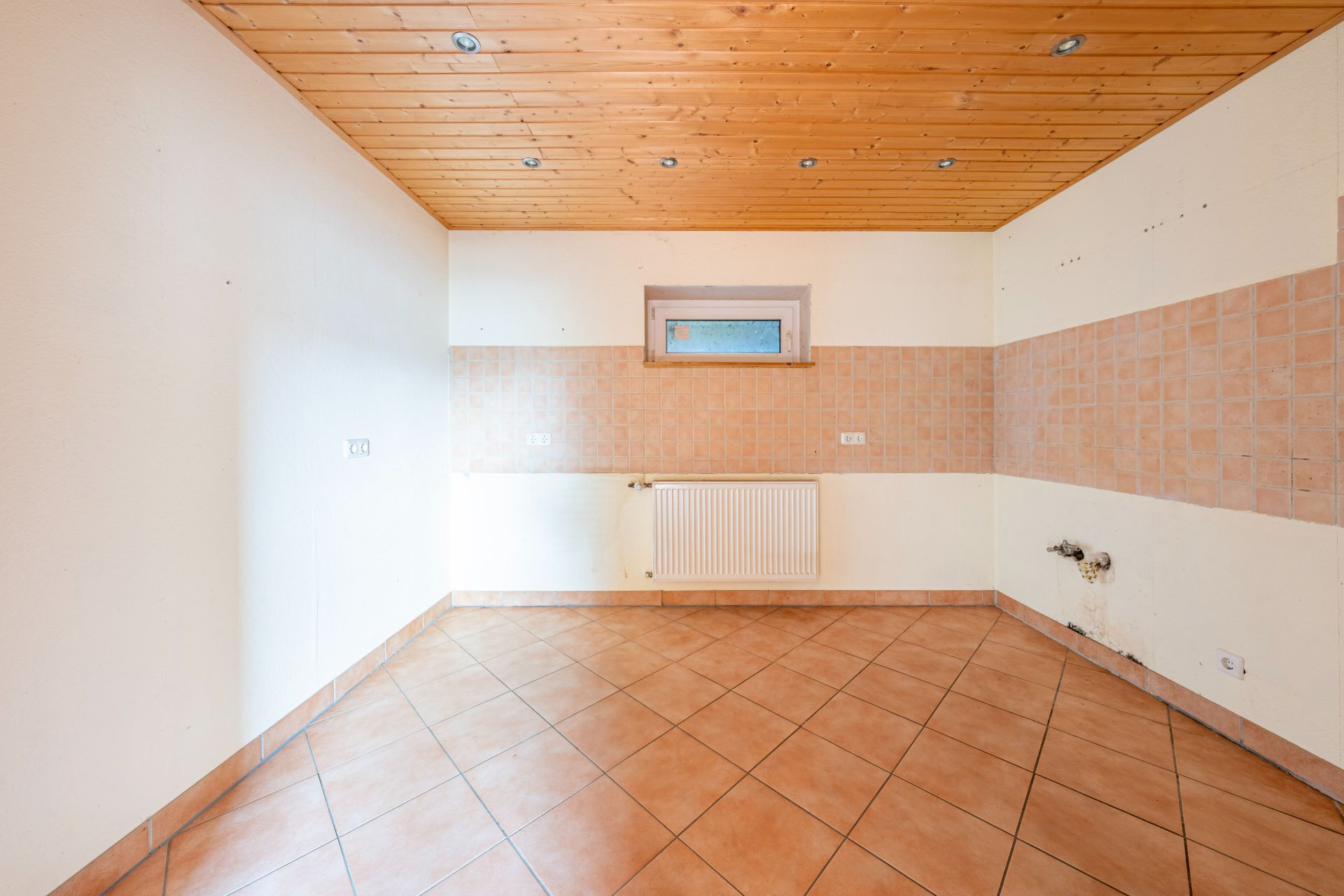Leerer Raum mit Fliesenboden, Holzdecke, kleinem Fenster und Fliesen an den Wänden.