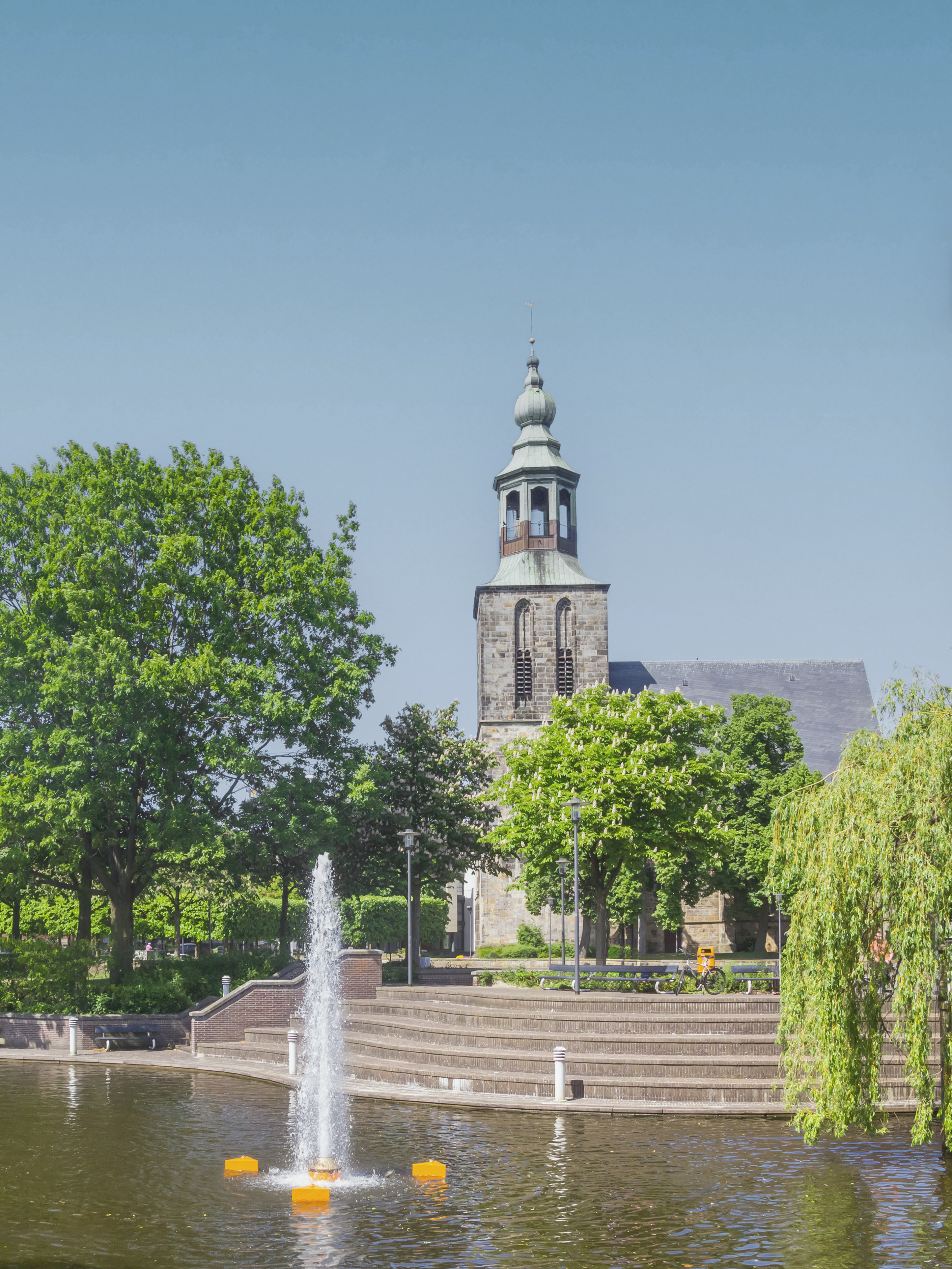Brunnen in einem Teich vor einer Kirche mit Bäumen und Treppen im Hintergrund.