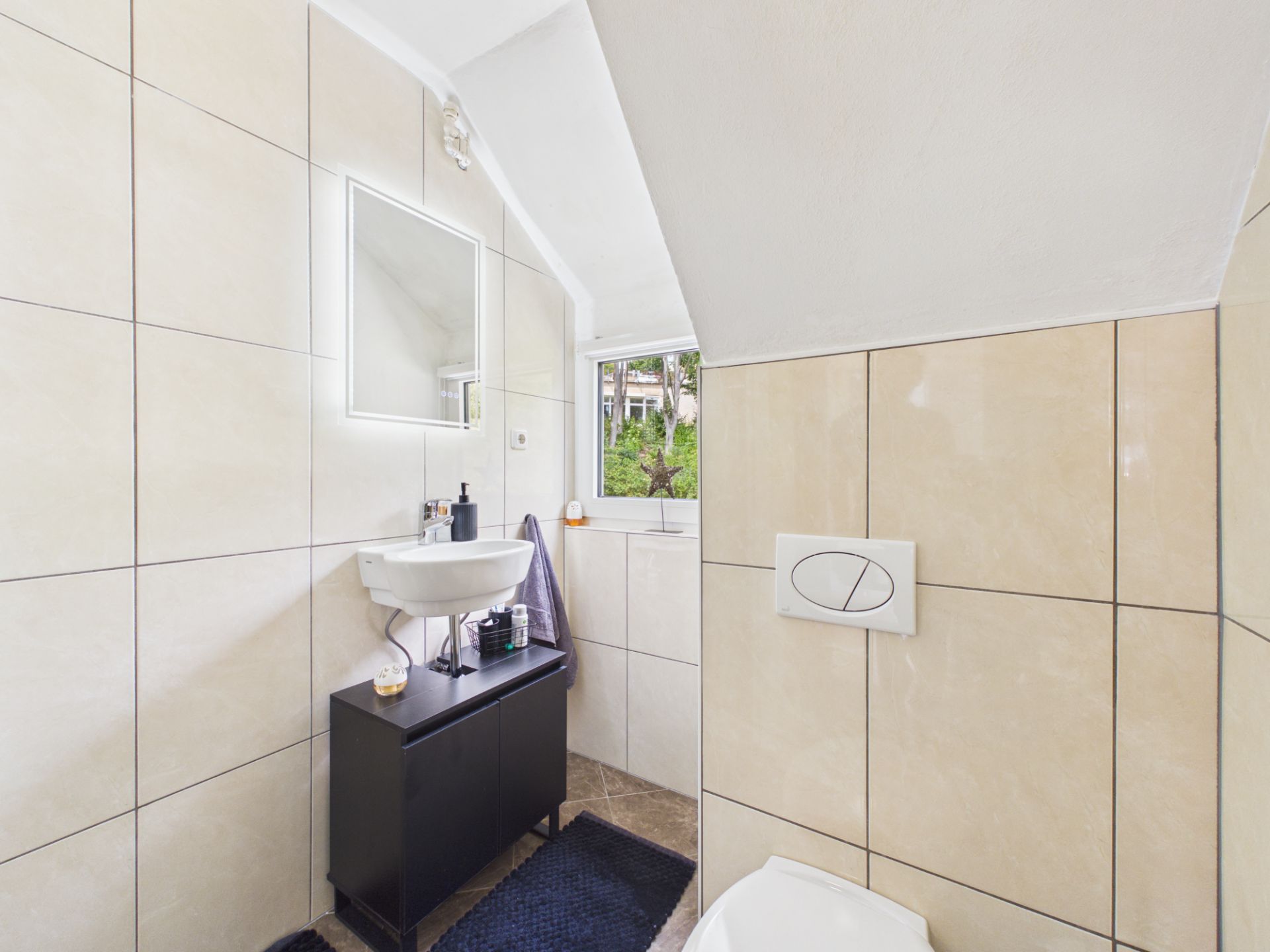 Modernes Bad mit Waschbecken, beleuchtetem Spiegel, WC und kleinem Fenster unter einer Dachschräge.