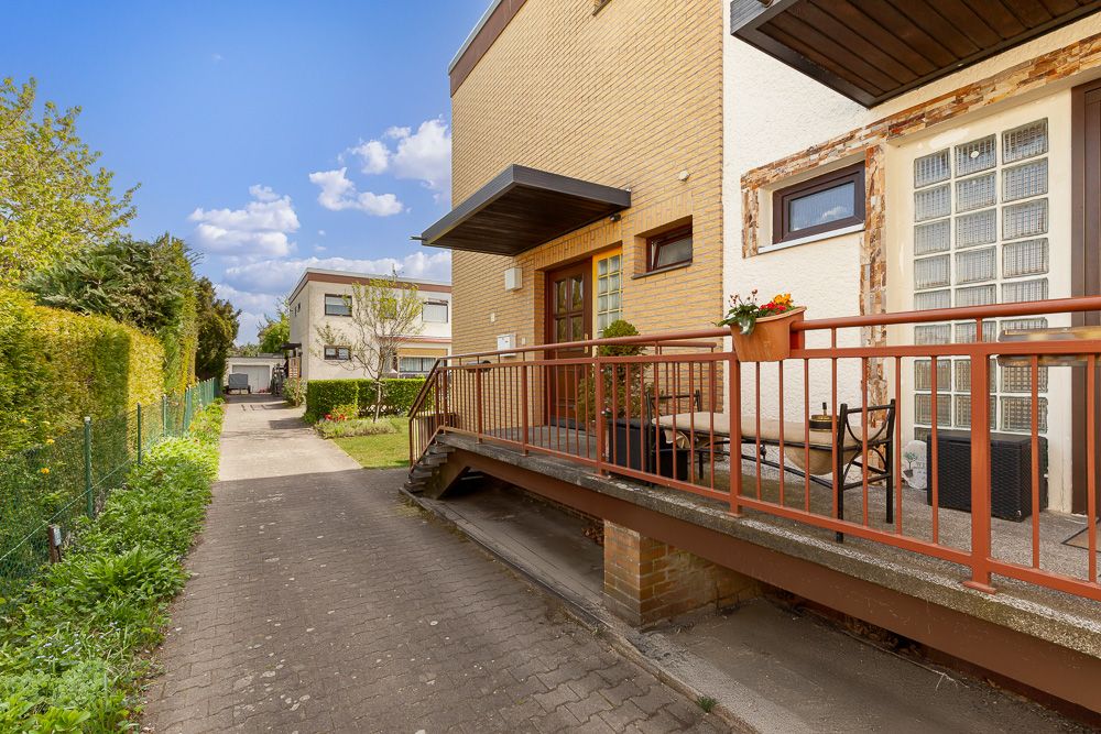 Vorgarten mit gepflastertem Weg, Terrasse mit Geländer und Sitzgelegenheiten an einem Wohnhaus.