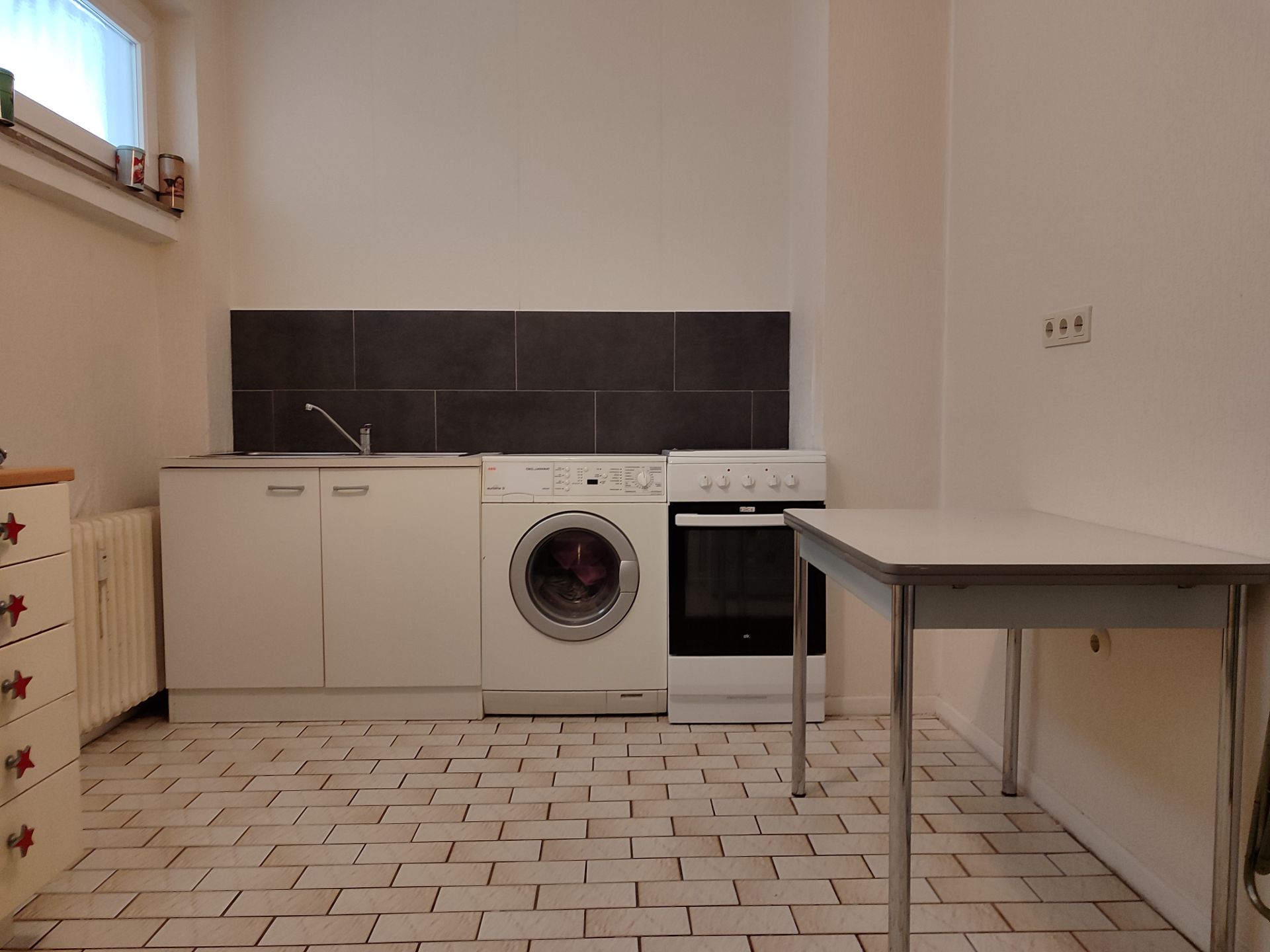Kleine Küche mit Waschmaschine, Herd, Spüle und Tisch auf gefliestem Boden.