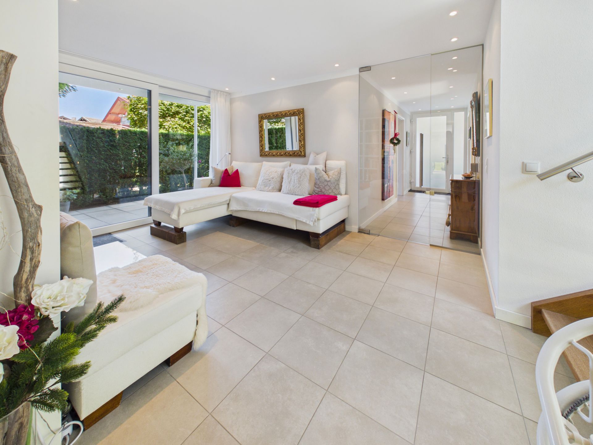 Helles Wohnzimmer mit weißem Ecksofa, Fliesenboden und großer Fensterfront zum Garten.