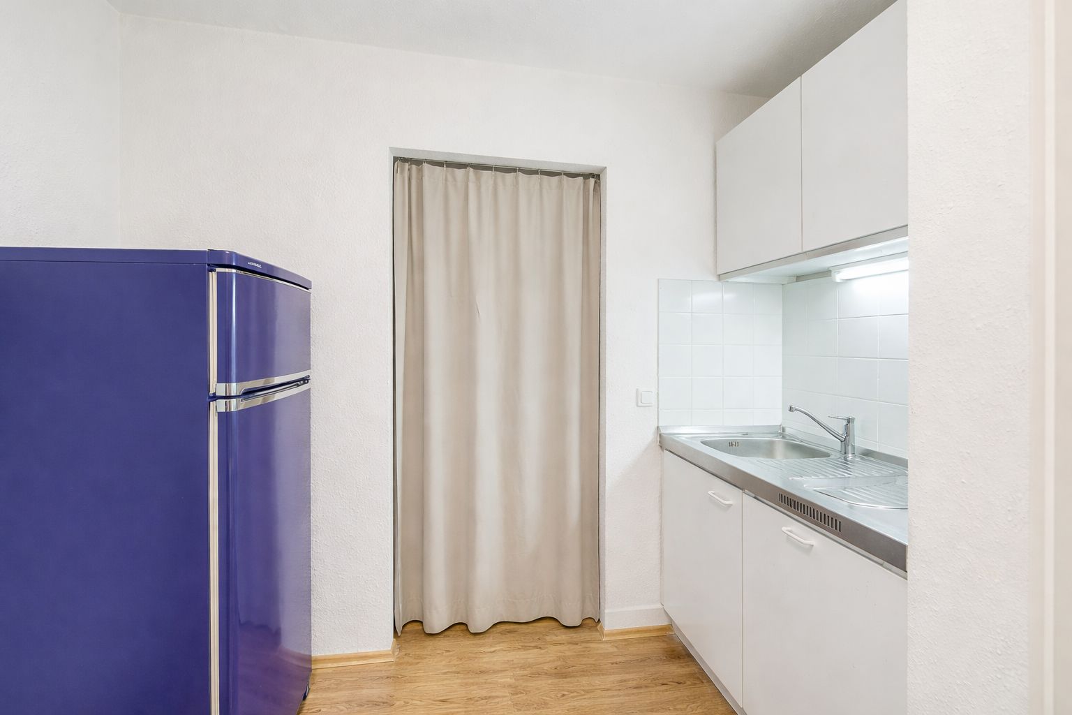 Kleine Küche mit blauem Kühlschrank, weißem Schrank und Vorhang vor der Tür.