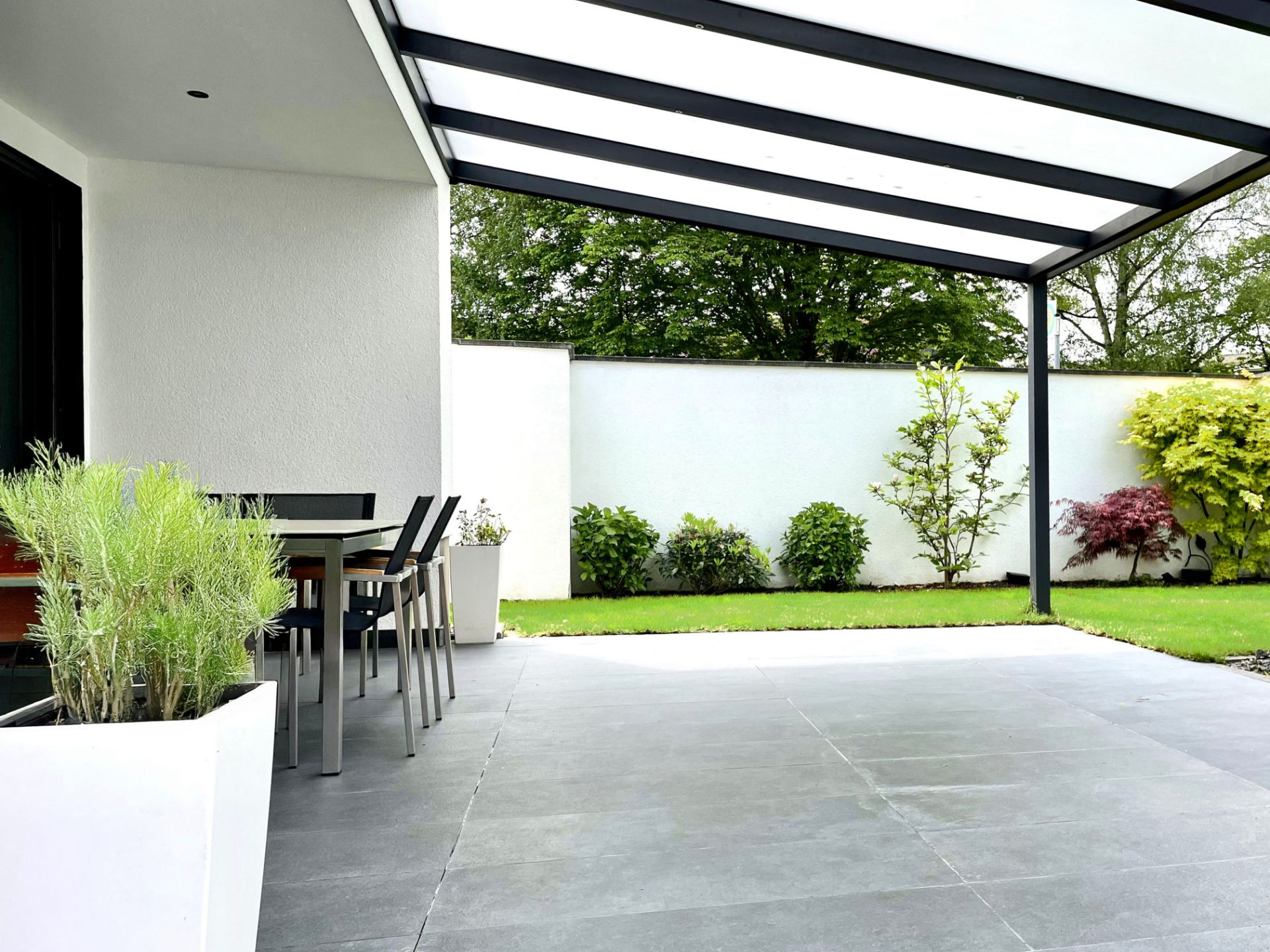 Überdachte Terrasse mit Esstisch und Stühlen, Blick auf Garten mit Rasen und Sträuchern.