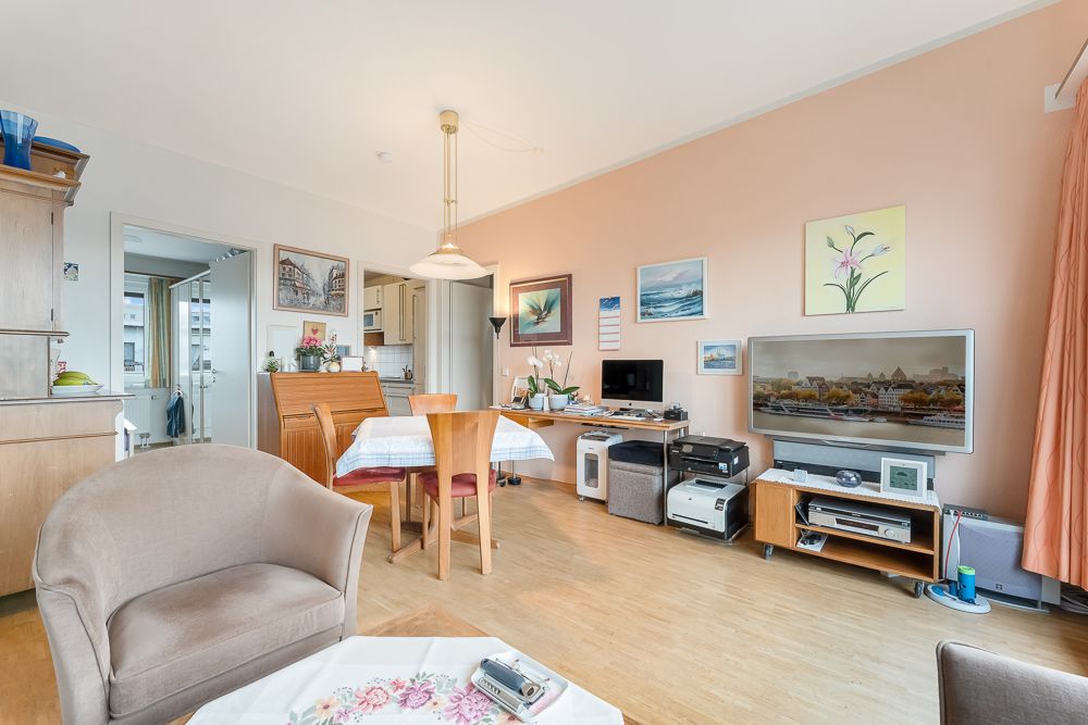 Helles Wohnzimmer mit Esstisch, Sesseln, Fernseher und mehreren Gemälden an der Wand.