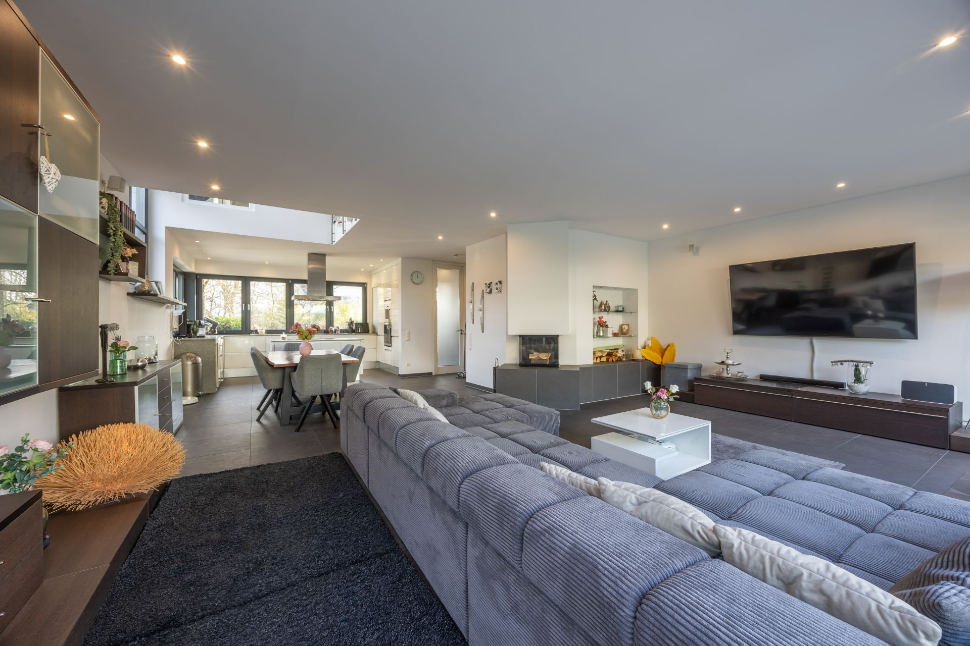 Modernes Wohnzimmer mit großer Eckcouch, Fernseher, Kamin und Essbereich im Hintergrund.