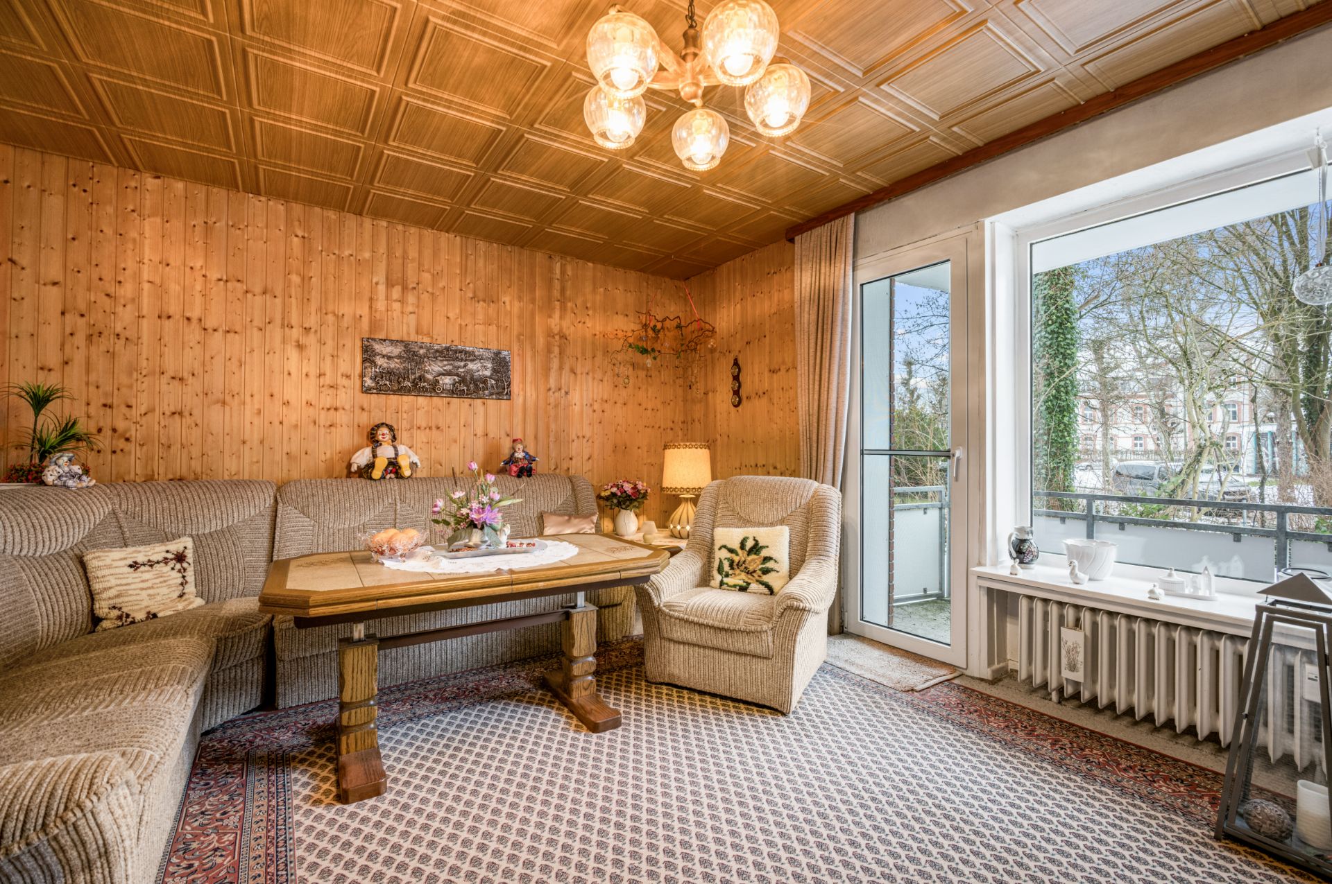 Gemütliches Wohnzimmer mit Ecksofa, Sessel, Holztisch und großem Fenster mit Blick nach draußen.