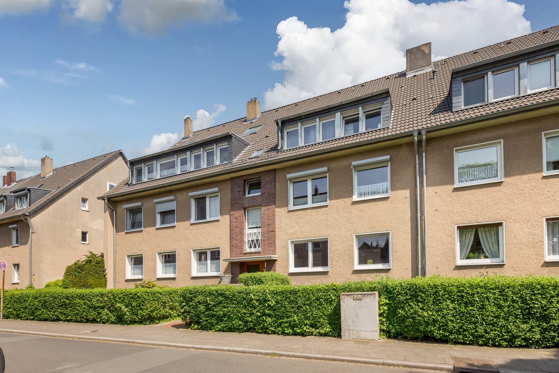 Mehrfamilienhaus mit drei Etagen, Satteldach und gepflegter Hecke an der Straße.