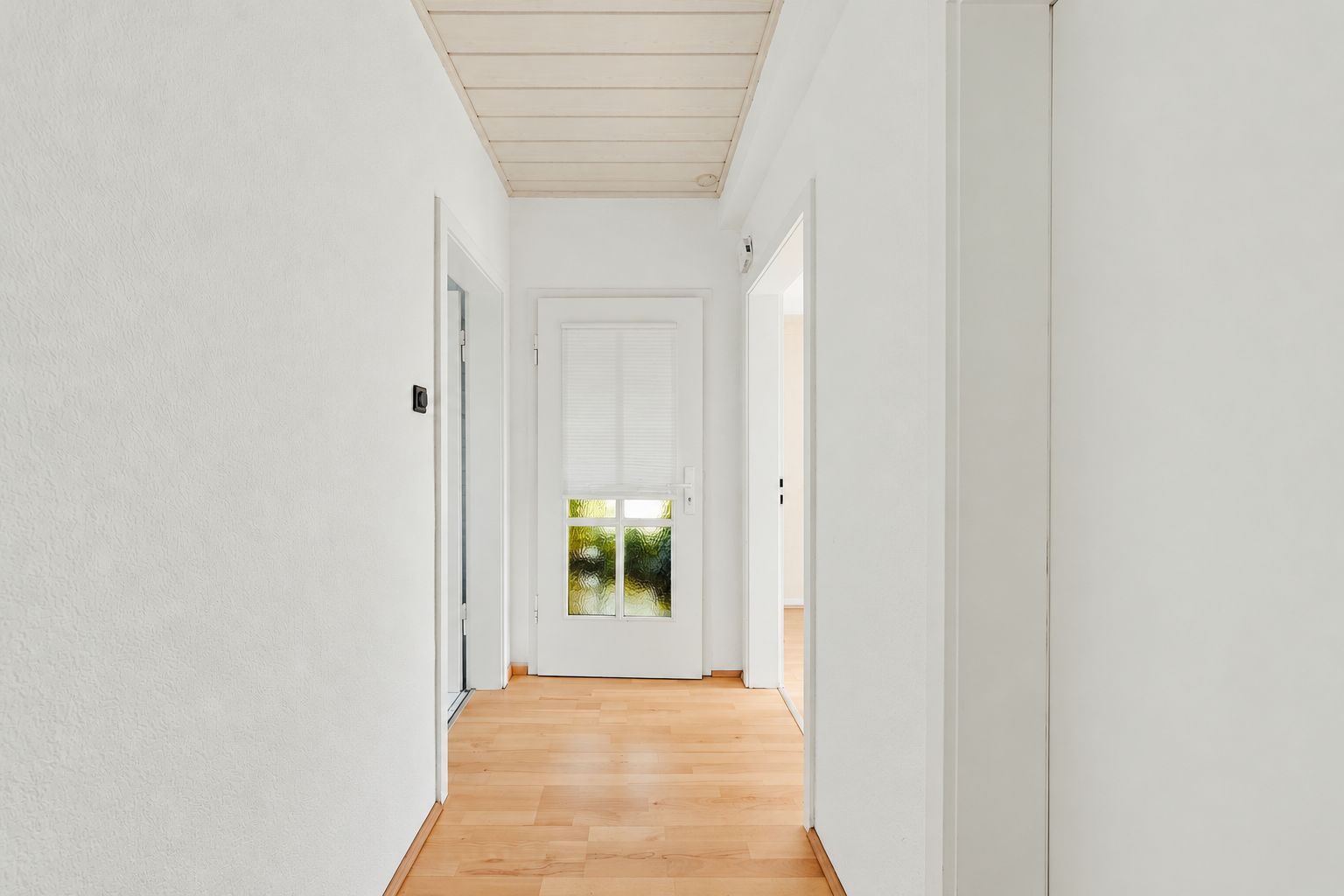 Heller Flur mit Holzboden, weißen Wänden und Tür mit Glasfeldern am Ende.