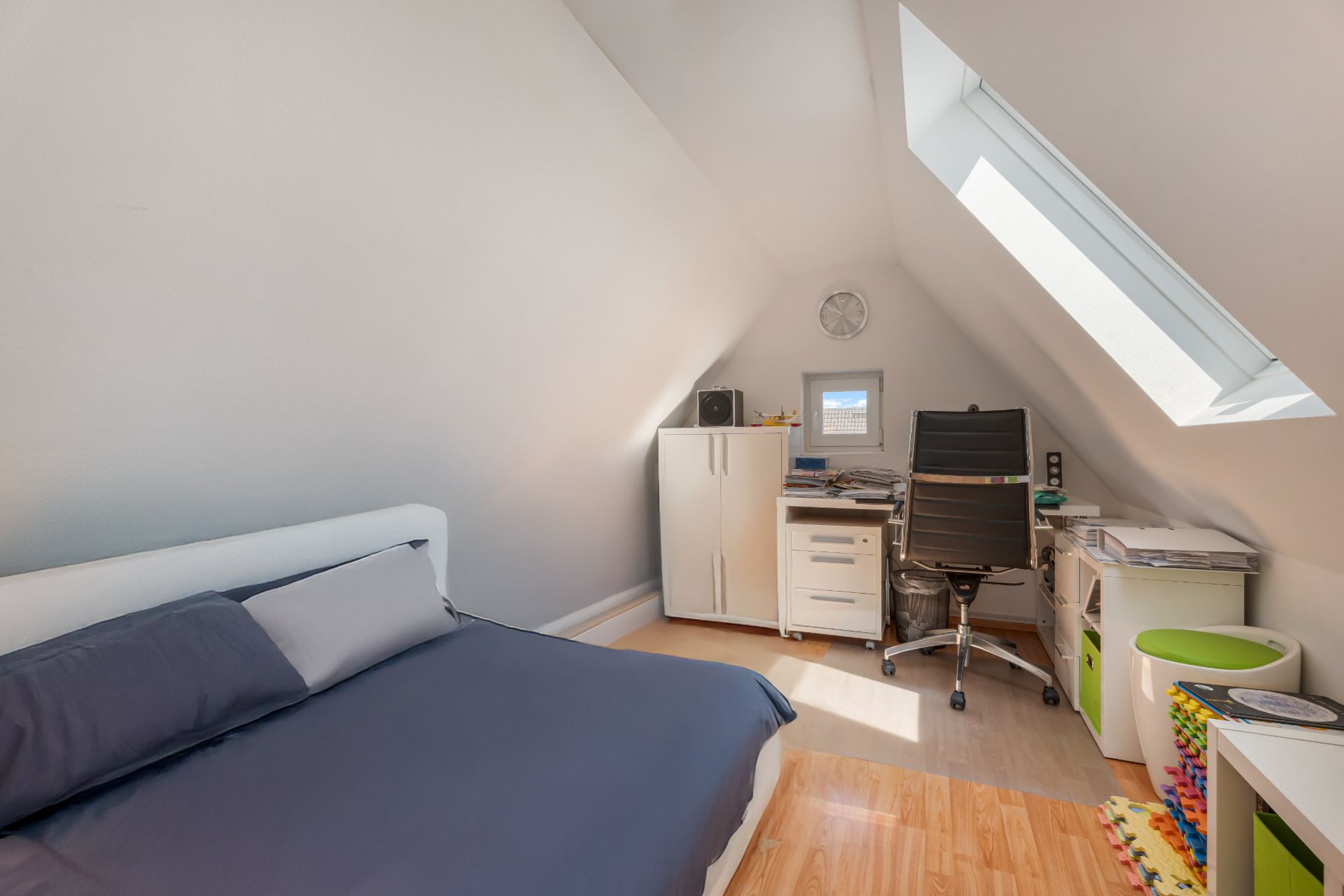 Dachzimmer mit Bett, Schreibtisch, Bürostuhl, Schränken und Dachfenster.