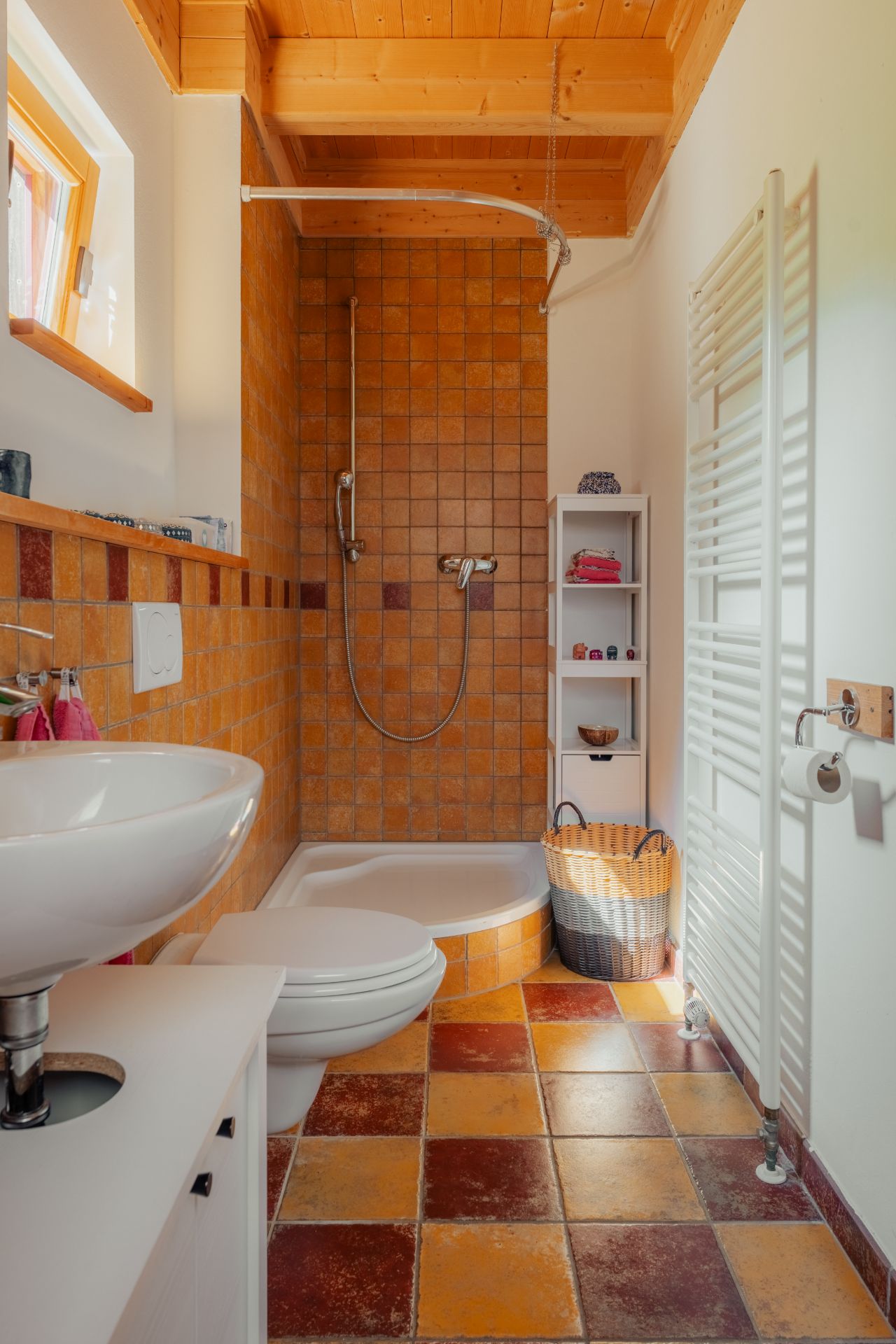 Kleines Badezimmer mit Dusche, Waschbecken, Toilette und Regal, gefliest in warmen Farben.