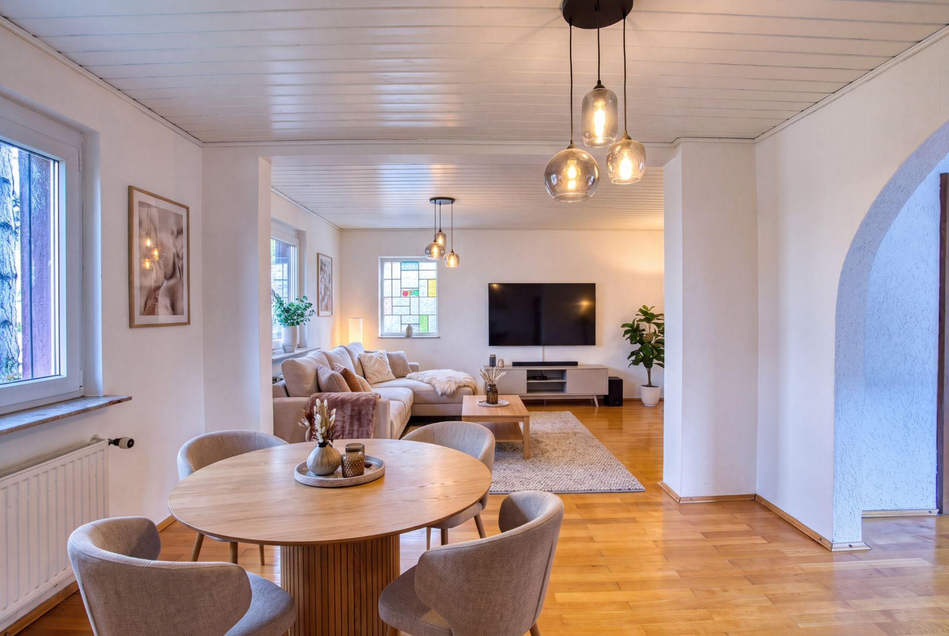 Modernes Wohnzimmer mit Sofa, Esstisch, Fernseher und großen Fenstern auf Holzfußboden.