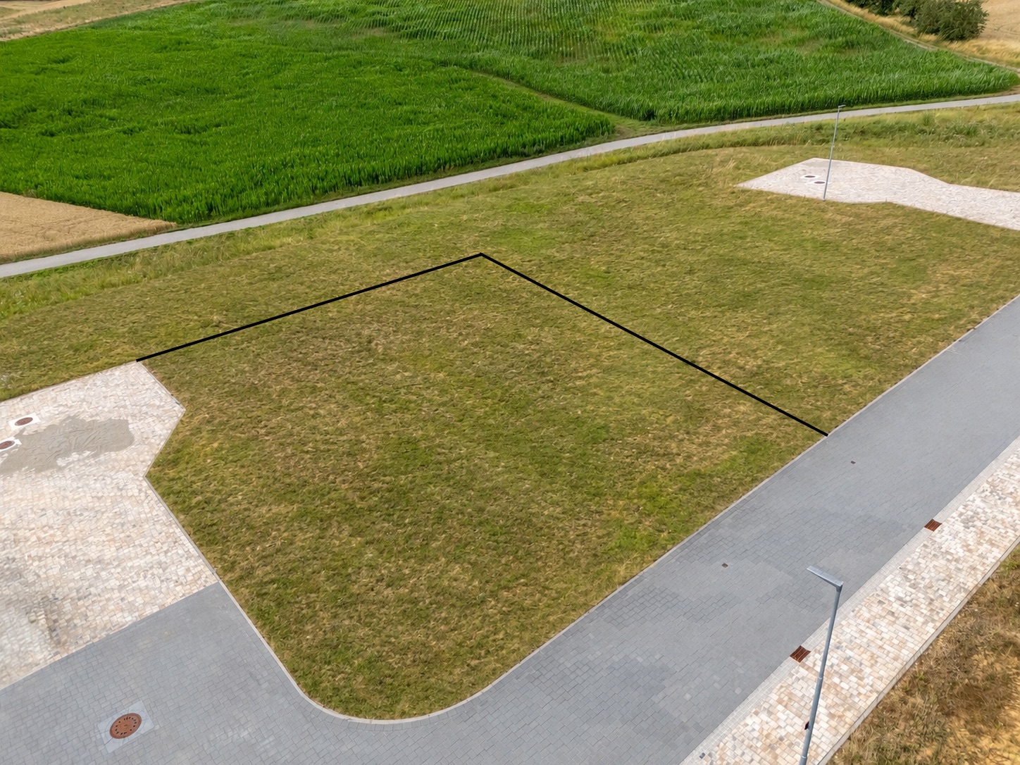 Leeres, rechteckiges Grundstück mit Rasen an einer gepflasterten Straße, im Hintergrund grüne Felder.