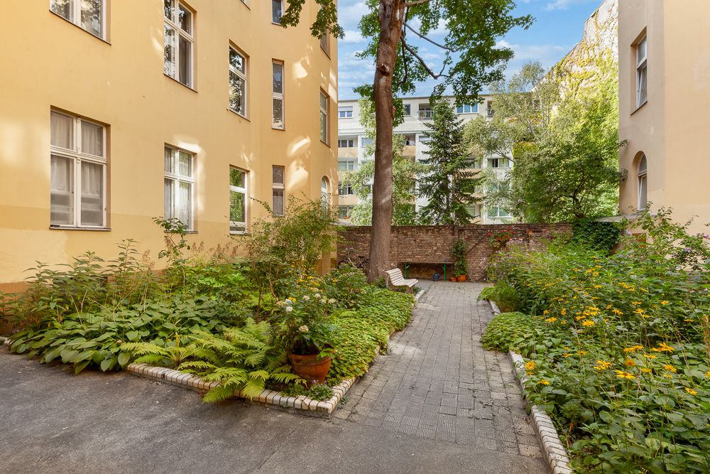 Innenhof mit bepflanzten Beeten, Pflasterweg, Bänken und gelbem Wohngebäude.