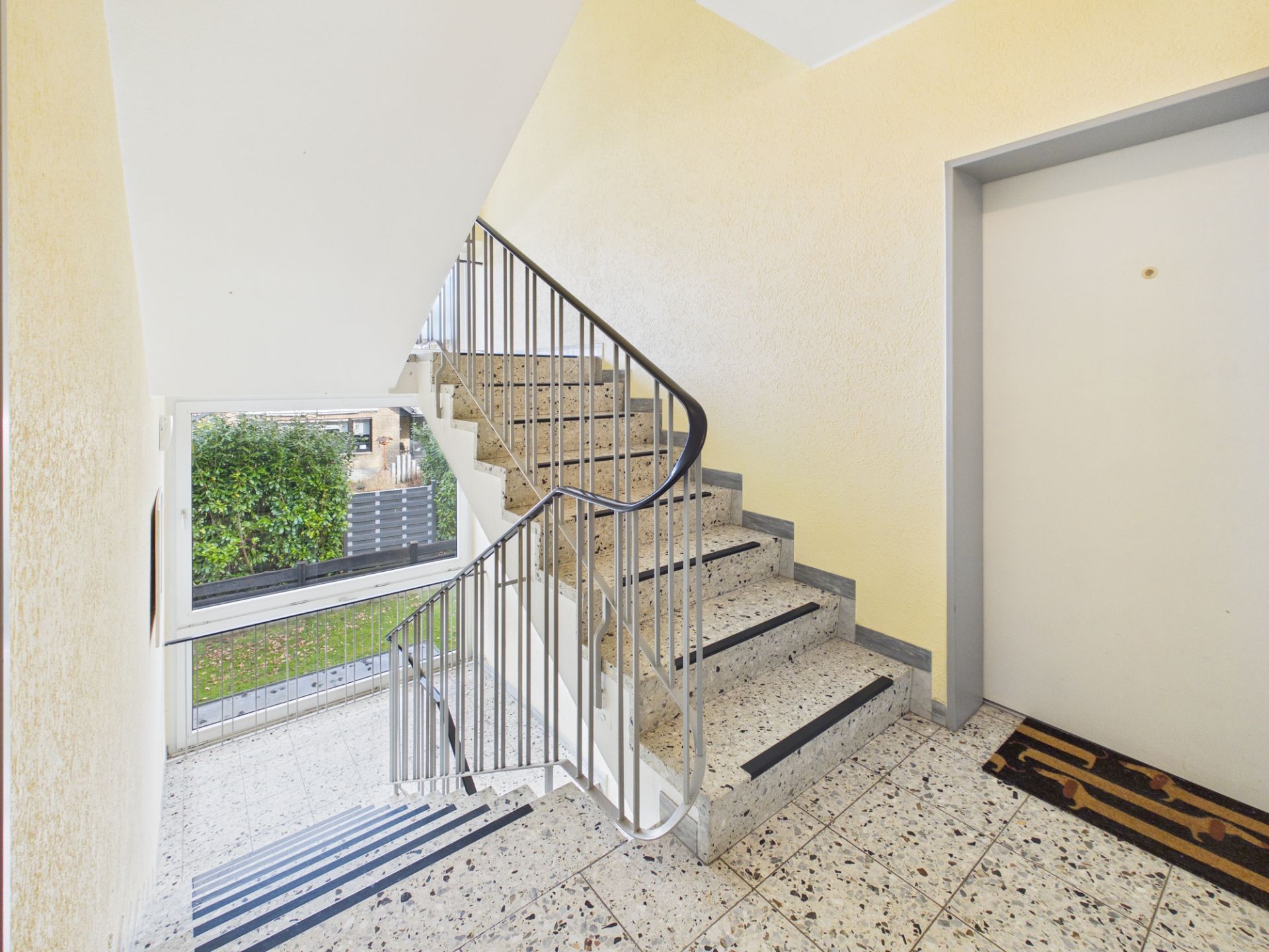 Helles Treppenhaus mit Geländer, Fenster und einer Wohnungstür auf der rechten Seite.