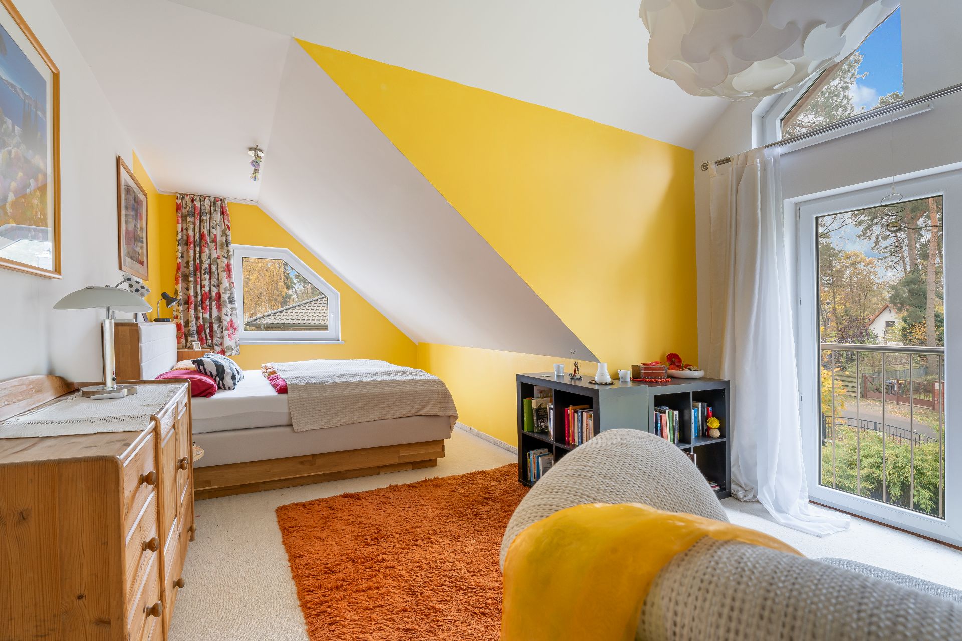 Helles Zimmer mit gelber Wand, Bett, Kommode, Bücherregal und großem Fenster mit Blick nach draußen.