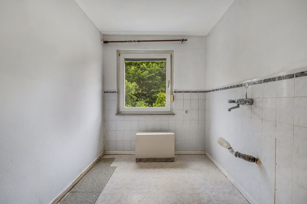 Leerer Raum mit weißen Wänden, gefliestem Bereich und Fenster mit Blick ins Grüne.