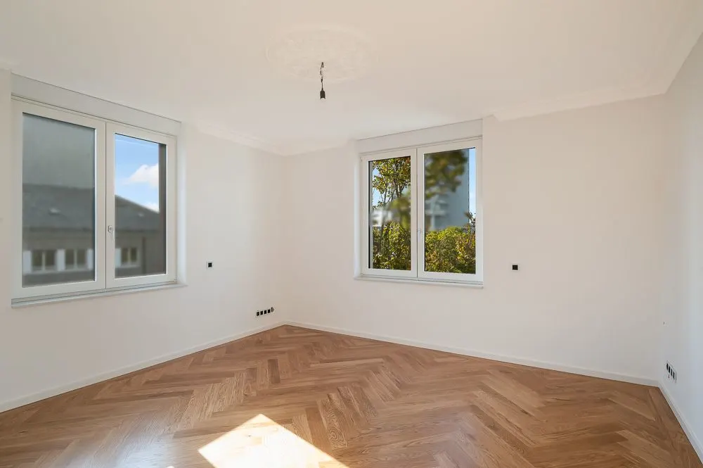 Leerer, heller Raum mit Parkettboden und zwei großen Fenstern.