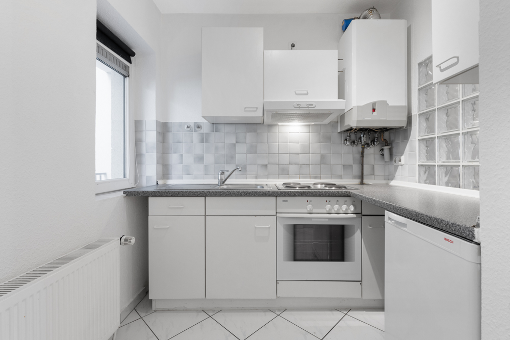 Helle, kleine Küche mit weißen Schränken, Herd, Backofen, Spüle und Fenster.