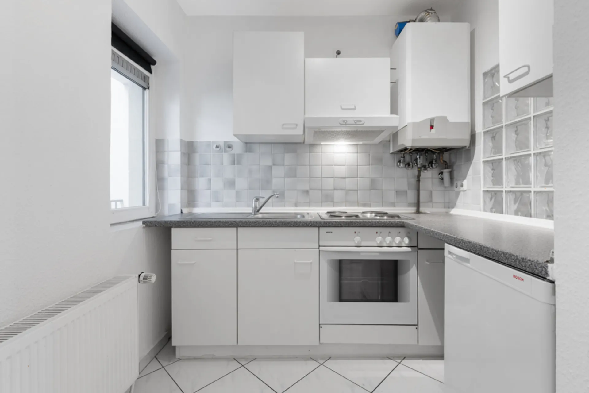 Helle, kleine Küche mit weißen Schränken, Herd, Backofen, Spüle und Fenster.