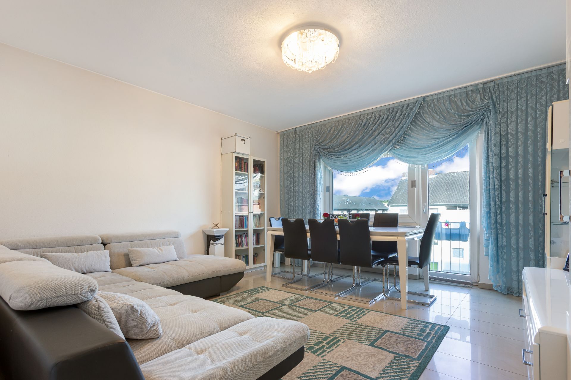 Wohnzimmer mit Ecksofa, Esstisch, acht Stühlen und großen Fenstern mit blauen Vorhängen.