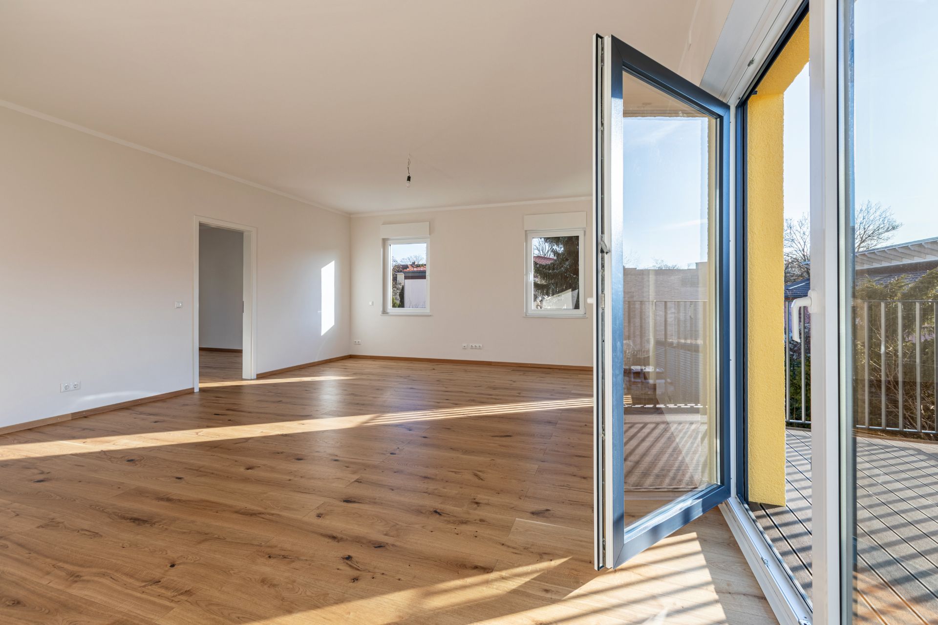 Helles, leeres Zimmer mit Holzboden, zwei Fenstern und geöffneter Glastür zu einem Balkon.