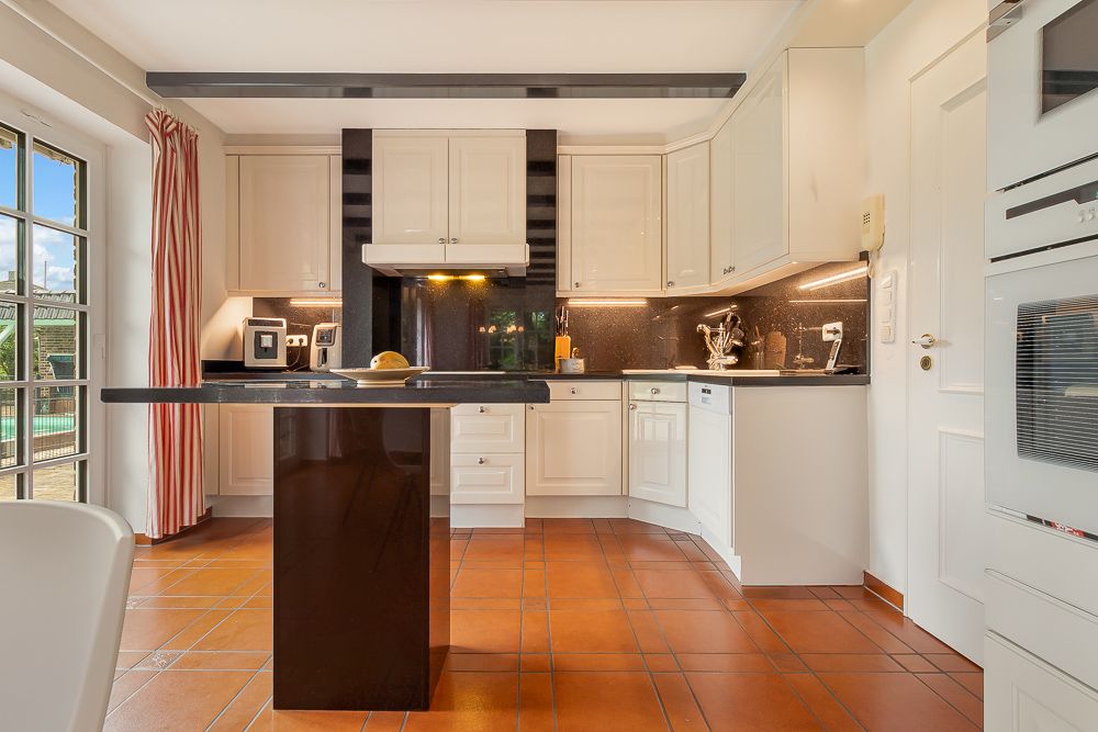 Moderne Küche mit weißen Schränken, schwarzer Arbeitsplatte und Kücheninsel, Terrakottafliesen am Boden.